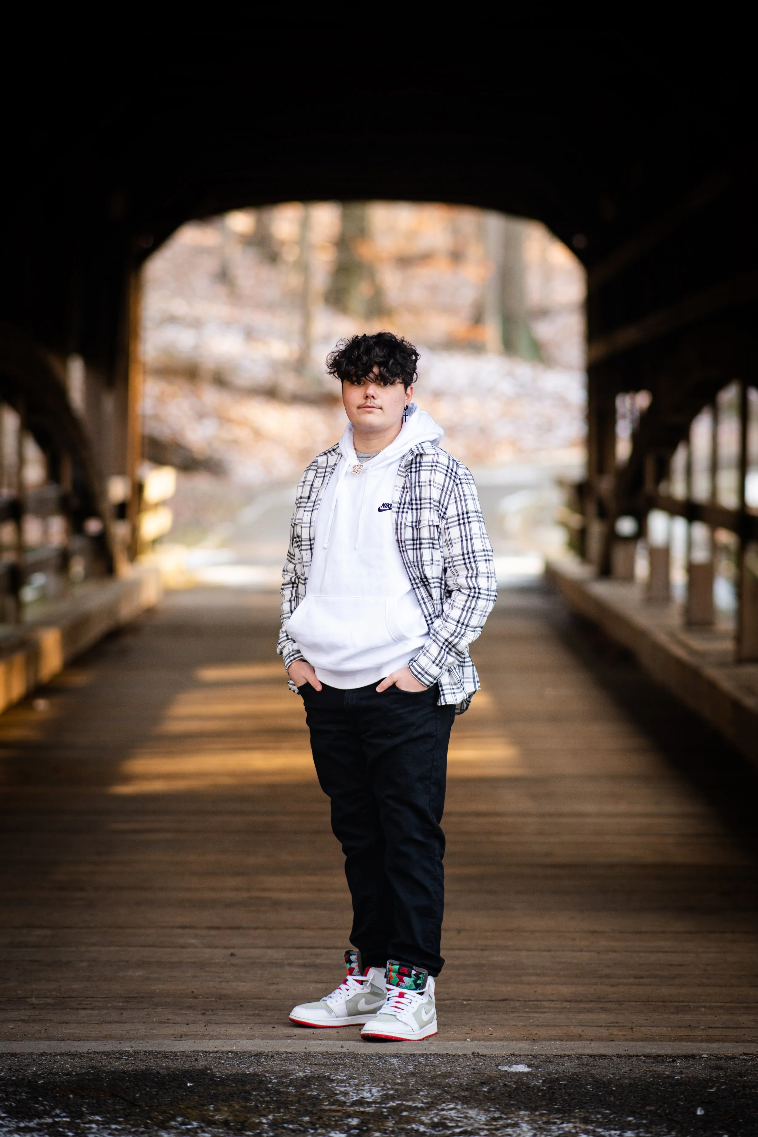 A young person with curly dark hair stands on a wooden bridge in a park during autumn, dressed in a white Nike hoodie, a plaid shirt, black pants, and white sneakers with colorful accents.