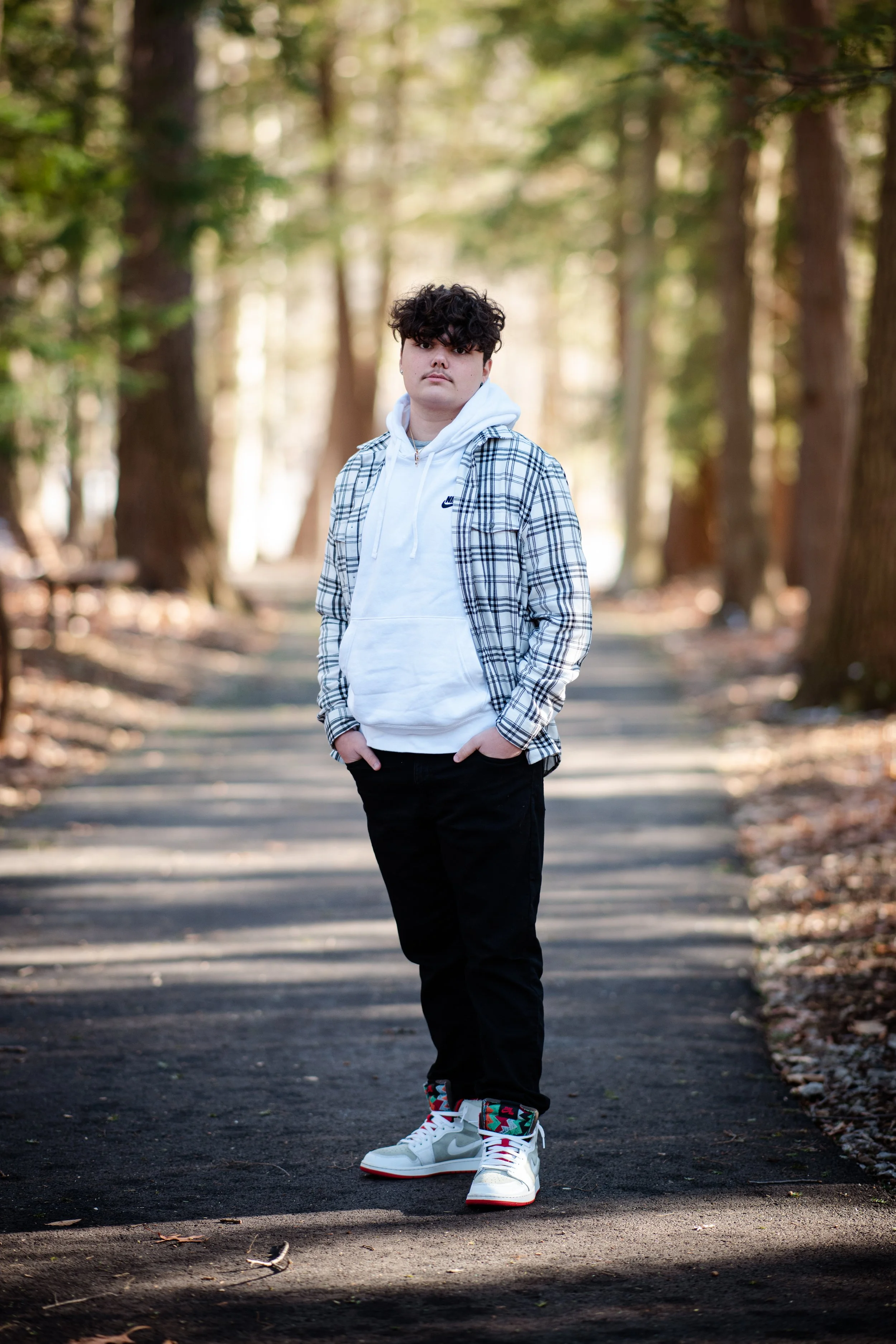 A young man standing on a forest trail during daytime, wearing a white hoodie, black pants, a plaid shirt, and colorful sneakers.