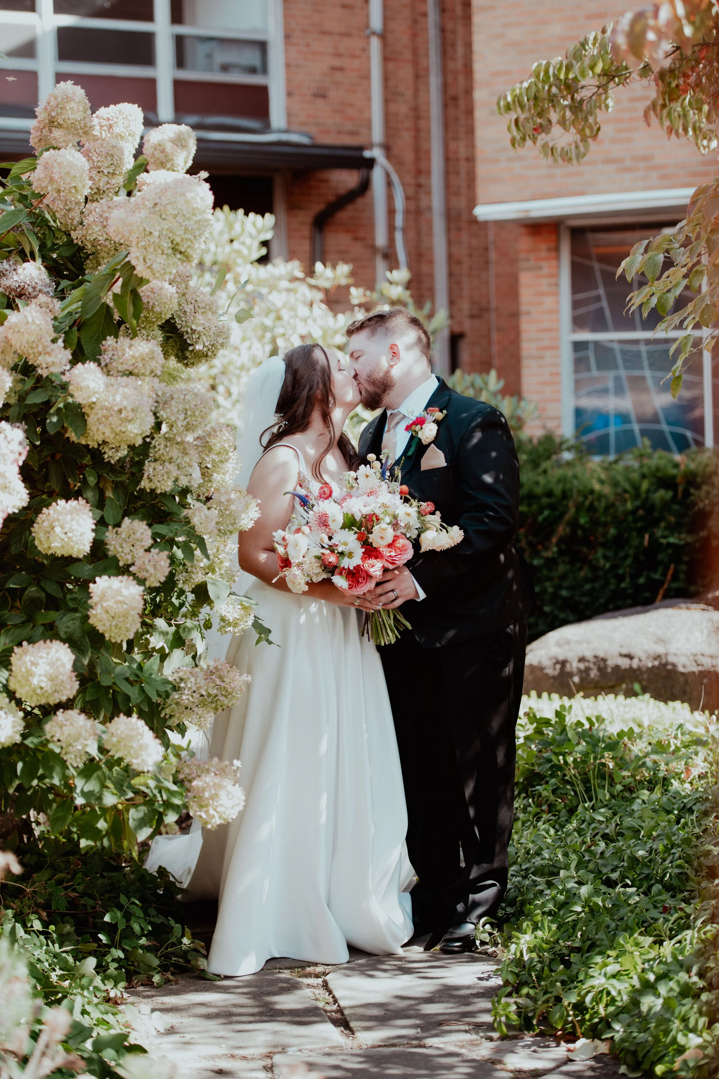 A bride and groom sharing a kiss outdoors, surrounded by blooming hydrangeas and garden greenery, with a brick building in the background.
