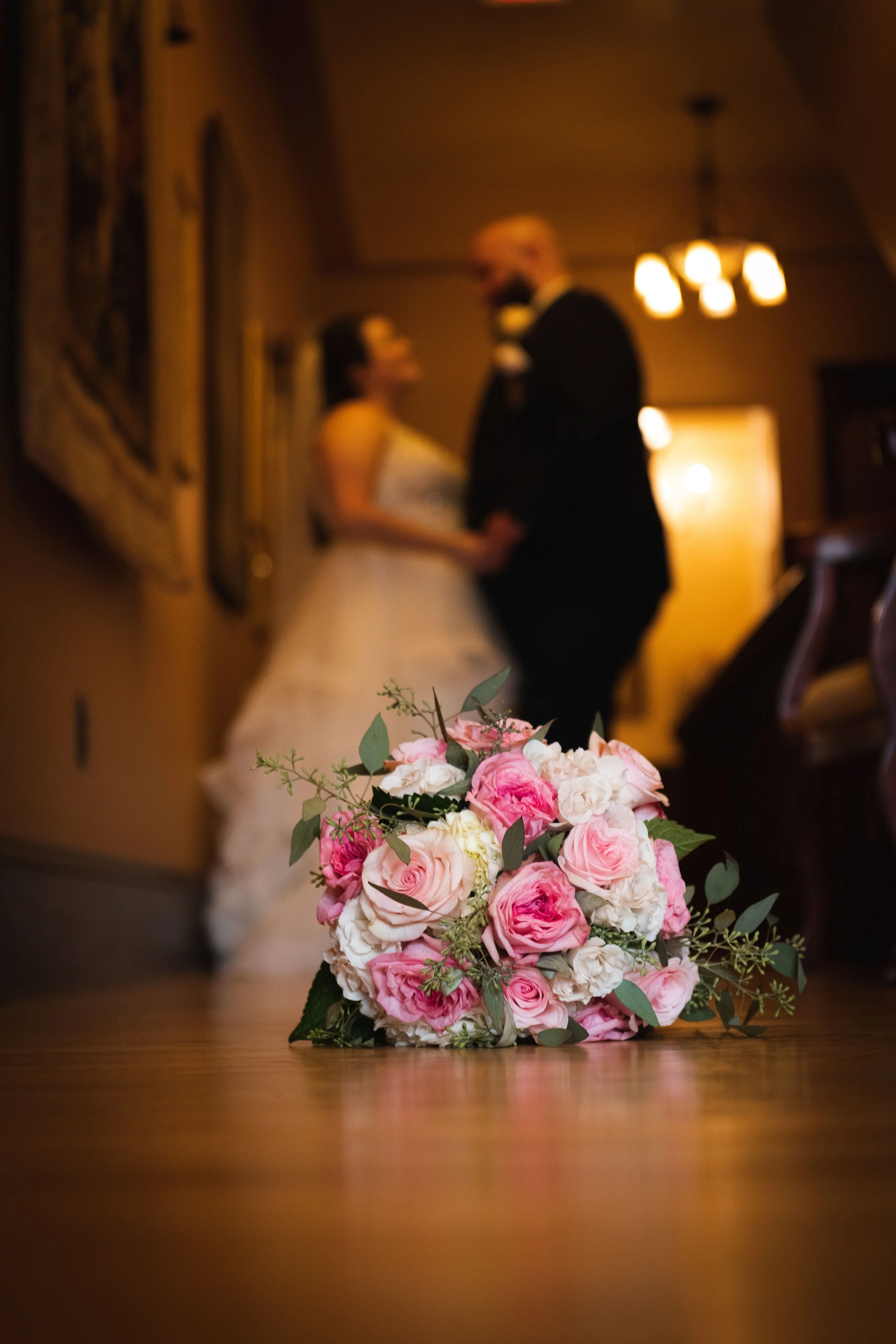Wedding bouquet of pink and white roses on a wooden floor with a blurred background of a bride and groom holding hands.