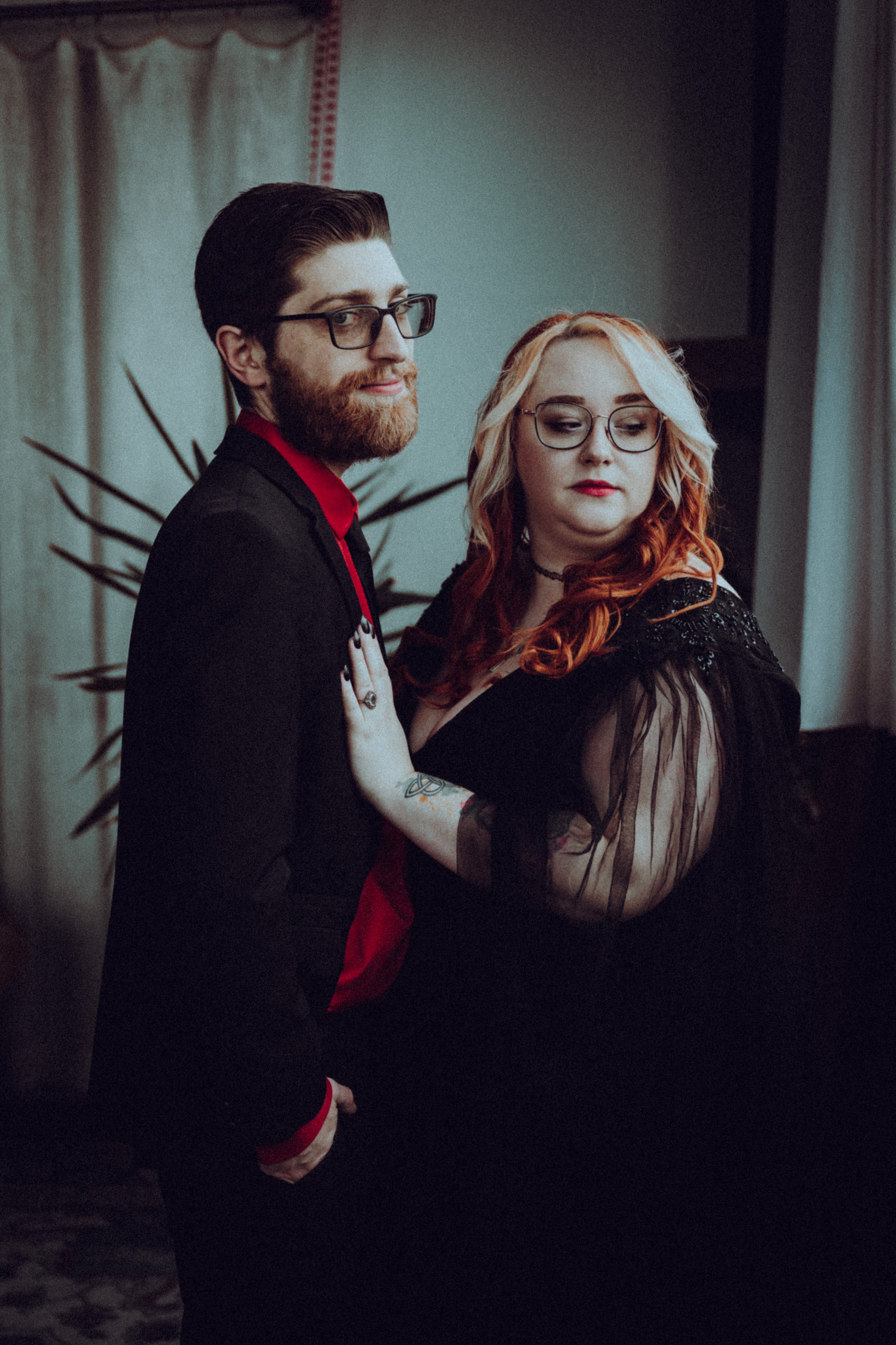A man with glasses and a beard wearing a black suit and red shirt, and a woman with blonde and red hair wearing glasses and a black dress with sheer sleeves, standing close together indoors.