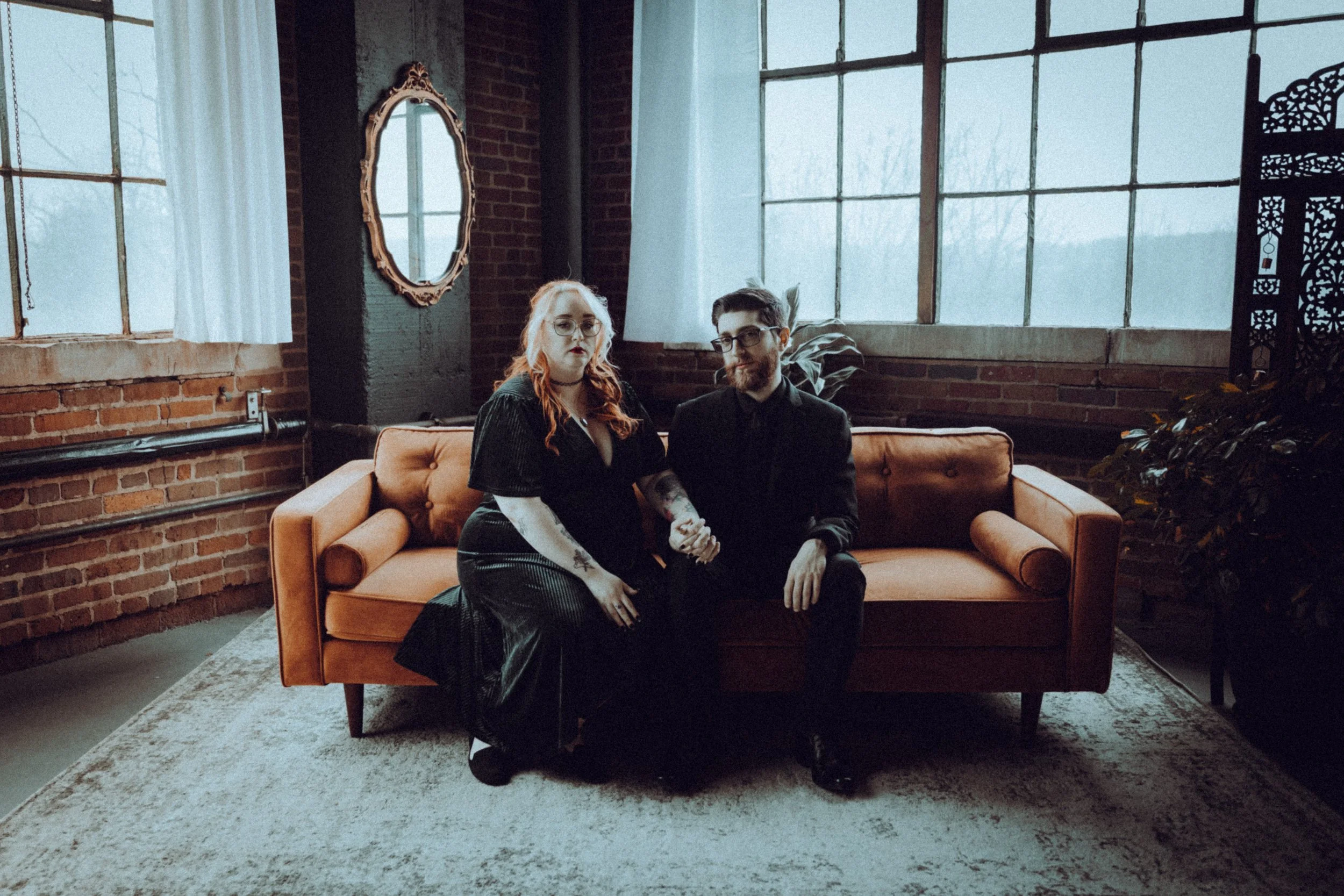 A woman with red hair and a man with glasses sitting on a brown velvet sofa in a room with brick walls and large industrial-style windows.