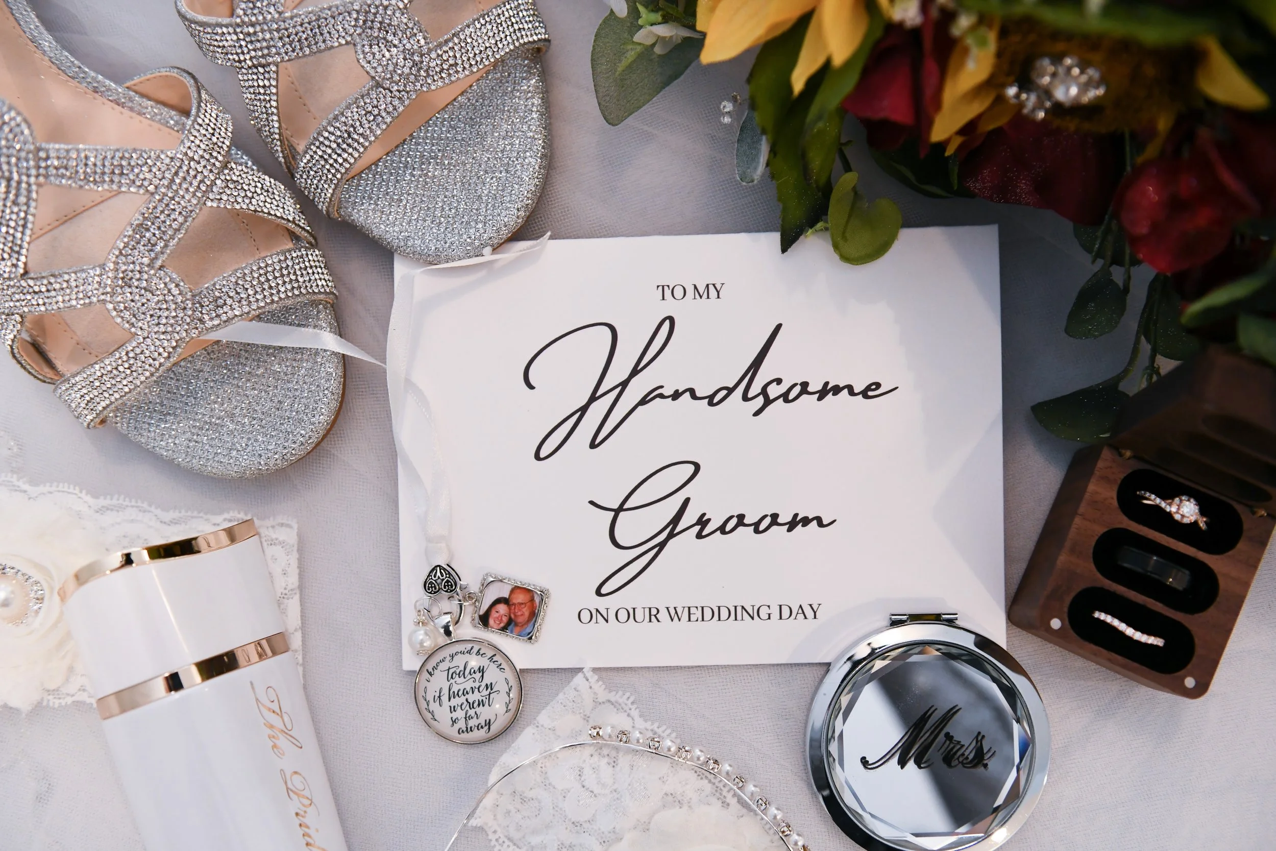 Wedding accessories including silver glitter high-heeled shoes, a wedding ring, a wedding vow book, a mirror with 'Mrs.' written on it, a bottle with 'Mrs. To Be' written on it, a small photo frame, and a bouquet of flowers.