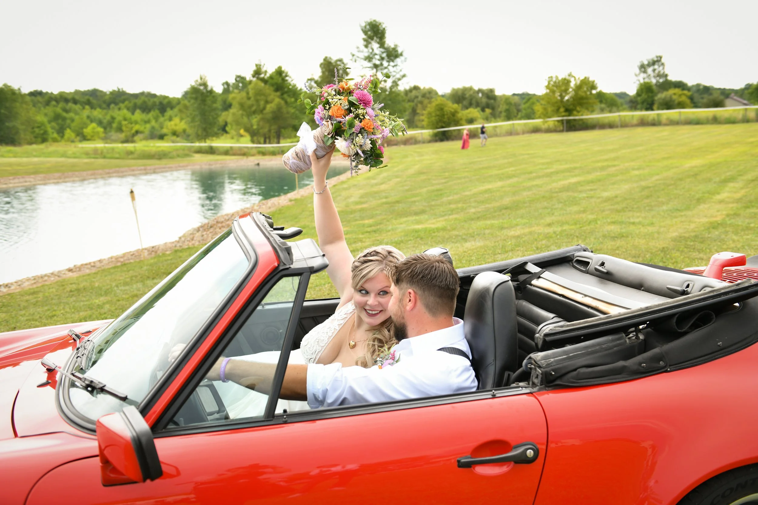 A newlywed couple in a red convertible car near a lake with green grass and trees in the background; the bride is holding a bouquet of flowers.