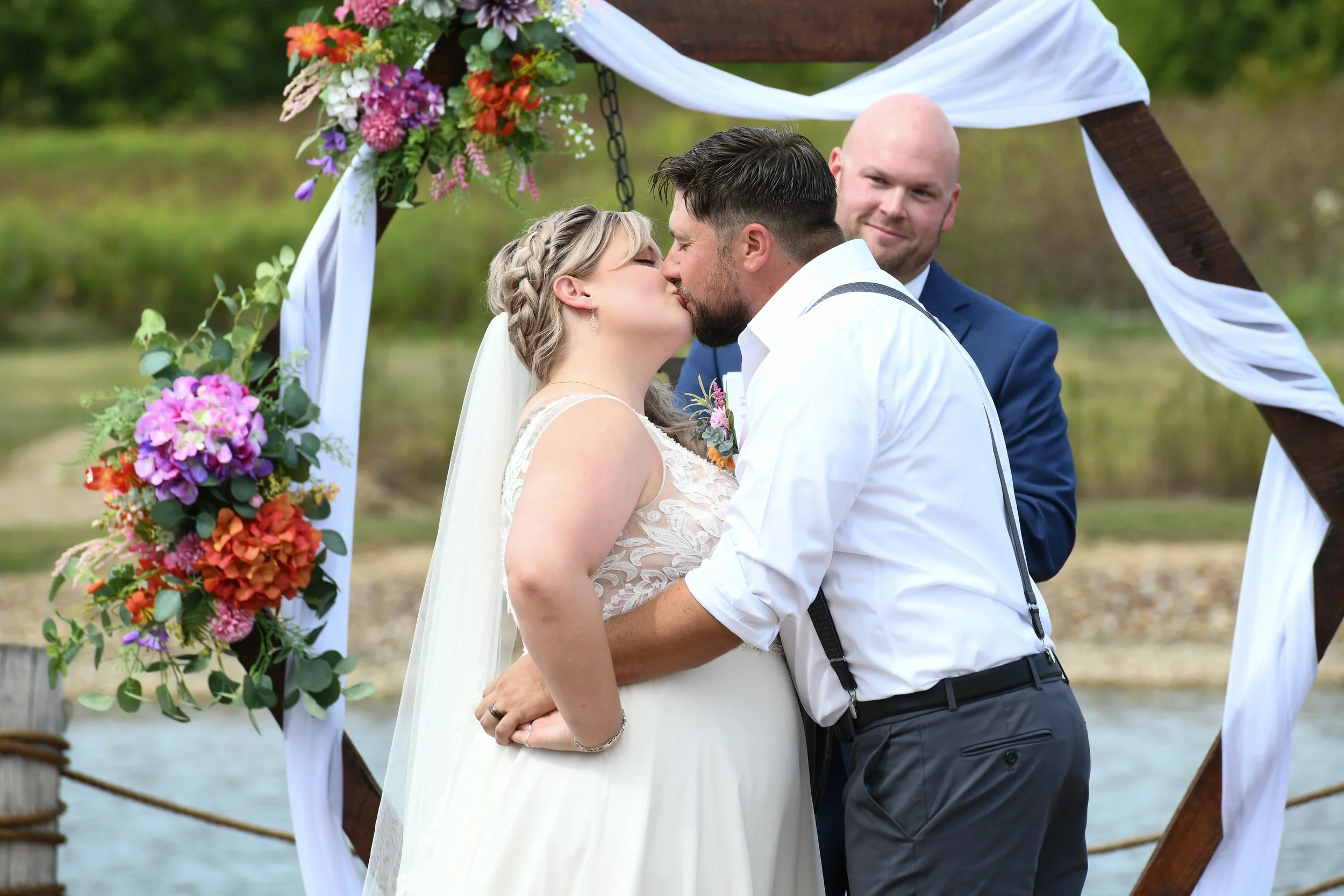 A bride and groom kiss during their outdoor wedding ceremony by a lake, with an officiant smiling in the background, under a wooden wedding arch decorated with colorful flowers and white drapes.