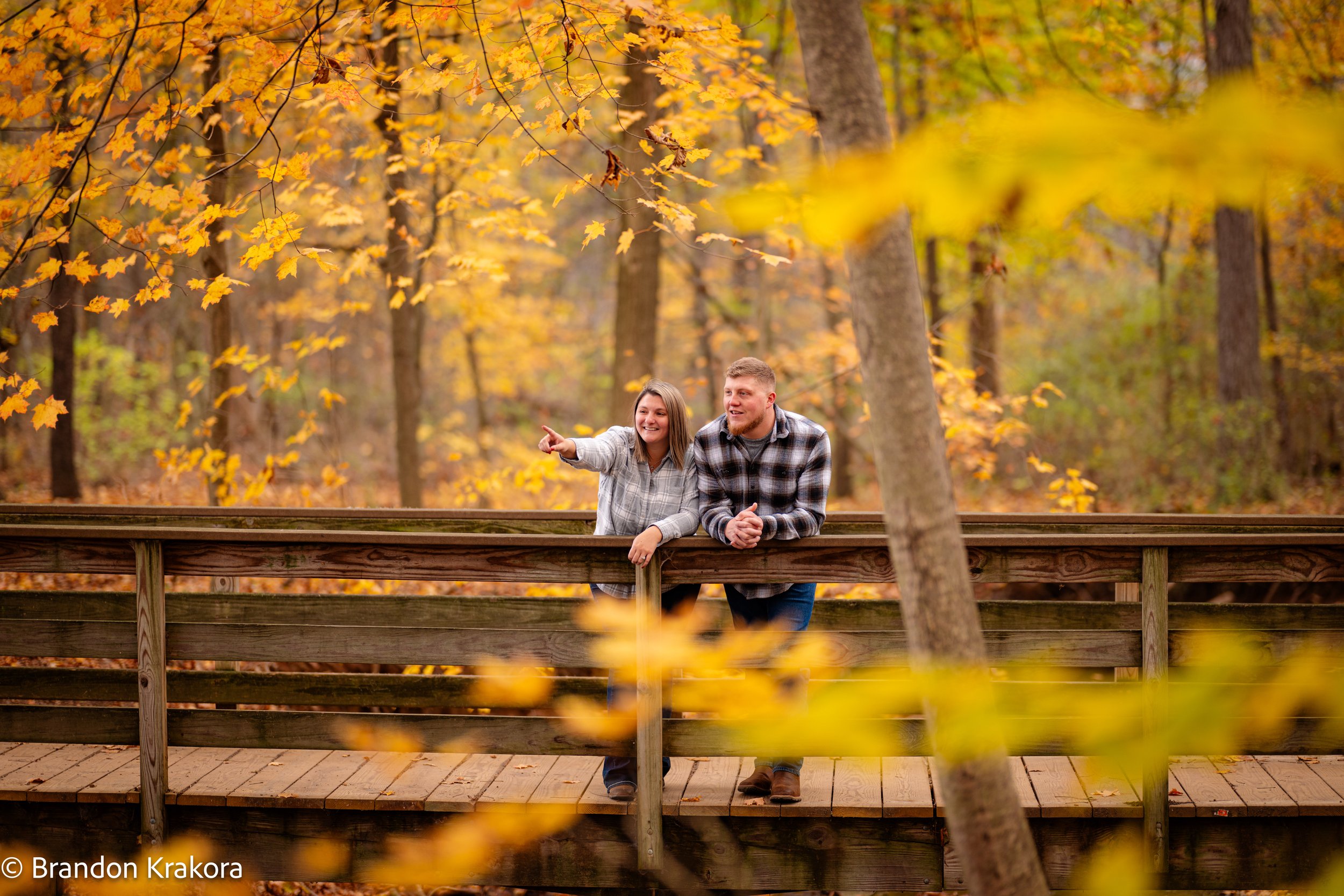 A young woman and man lean on a wooden railing on a bridge in a forest with autumn leaves, smiling and looking at something in the distance, with yellow and orange fall foliage around them.