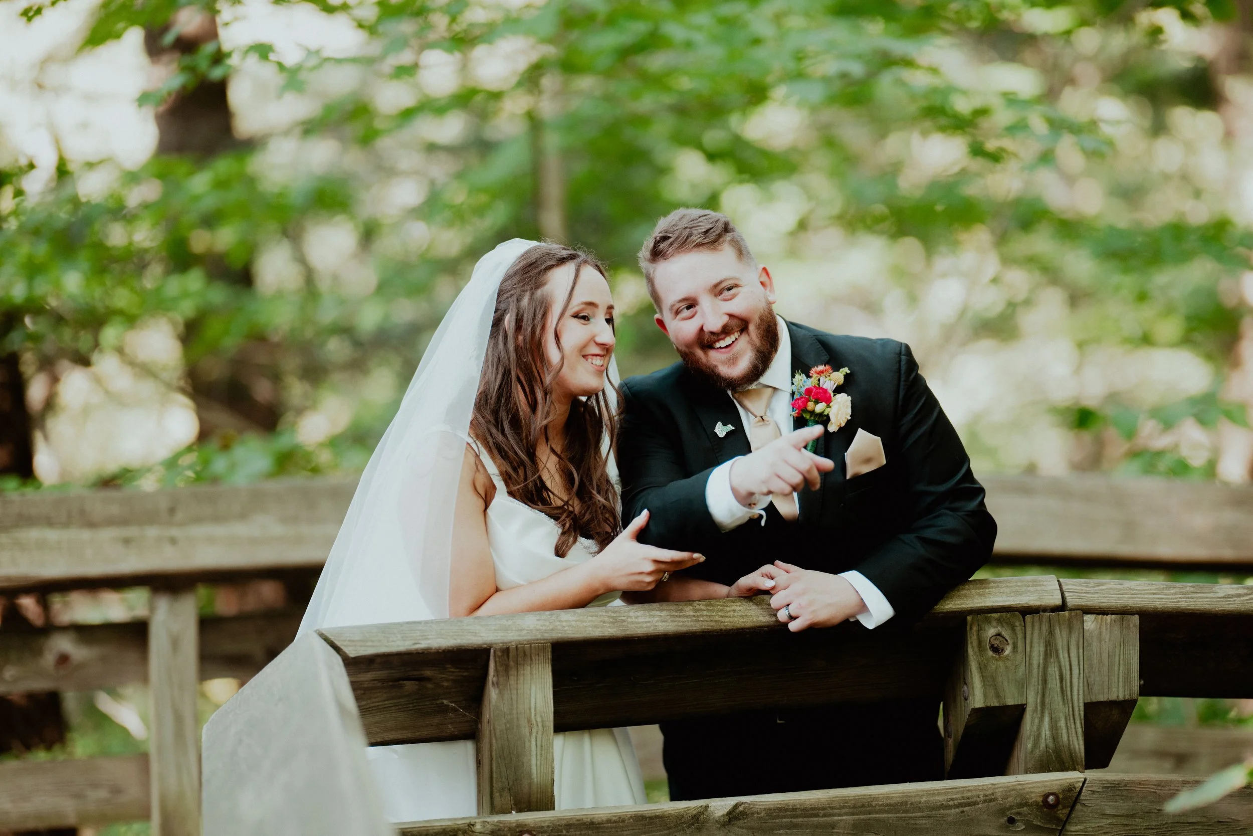 A bride and groom in wedding attire standing on a wooden bridge outdoors, smiling and looking at each other, with greenery in the background.