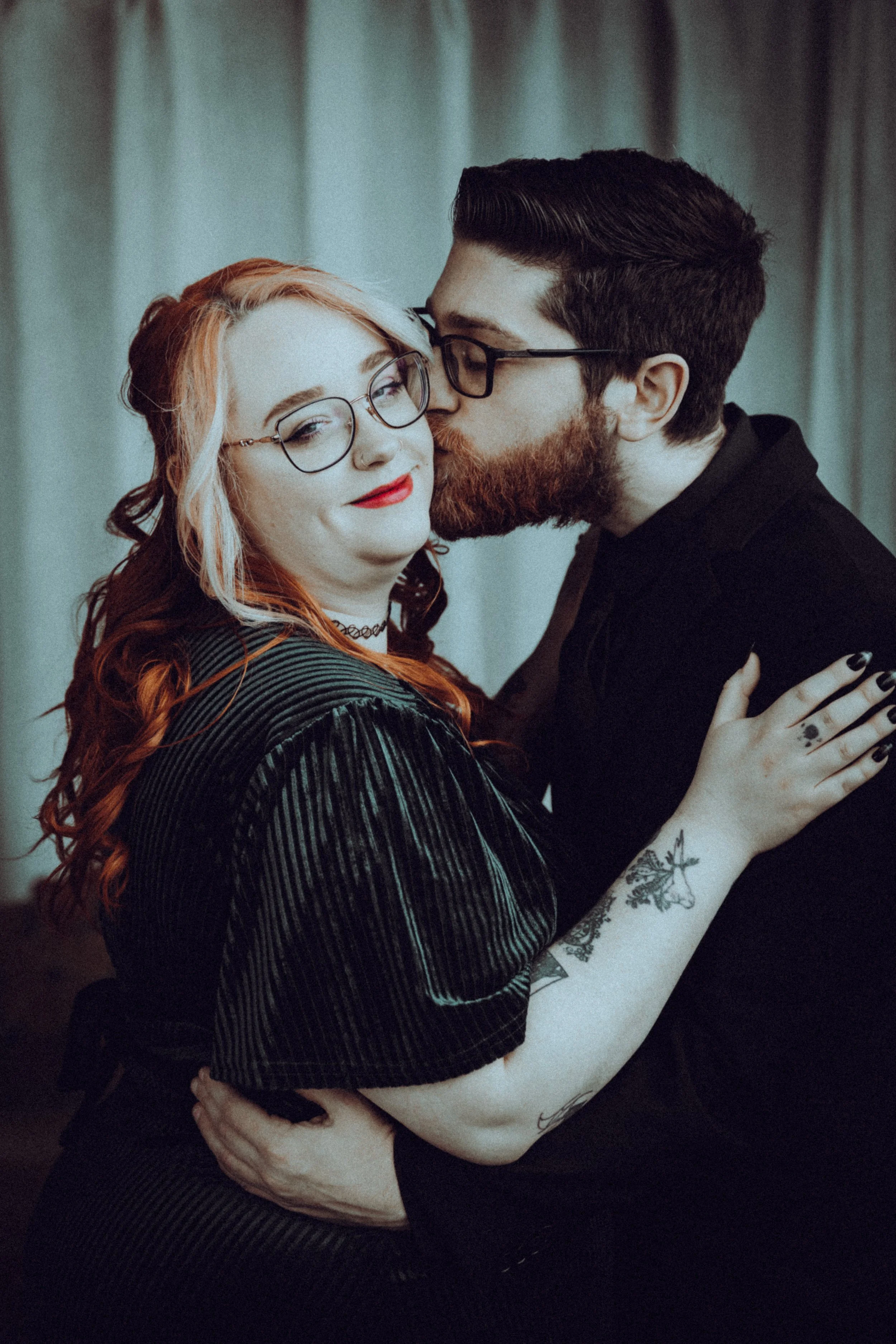A woman with red hair and glasses smiling as a man with dark hair and glasses kisses her on the cheek. They are holding each other closely, with the woman's arm around the man's shoulder, in front of a plain, light-colored curtain.