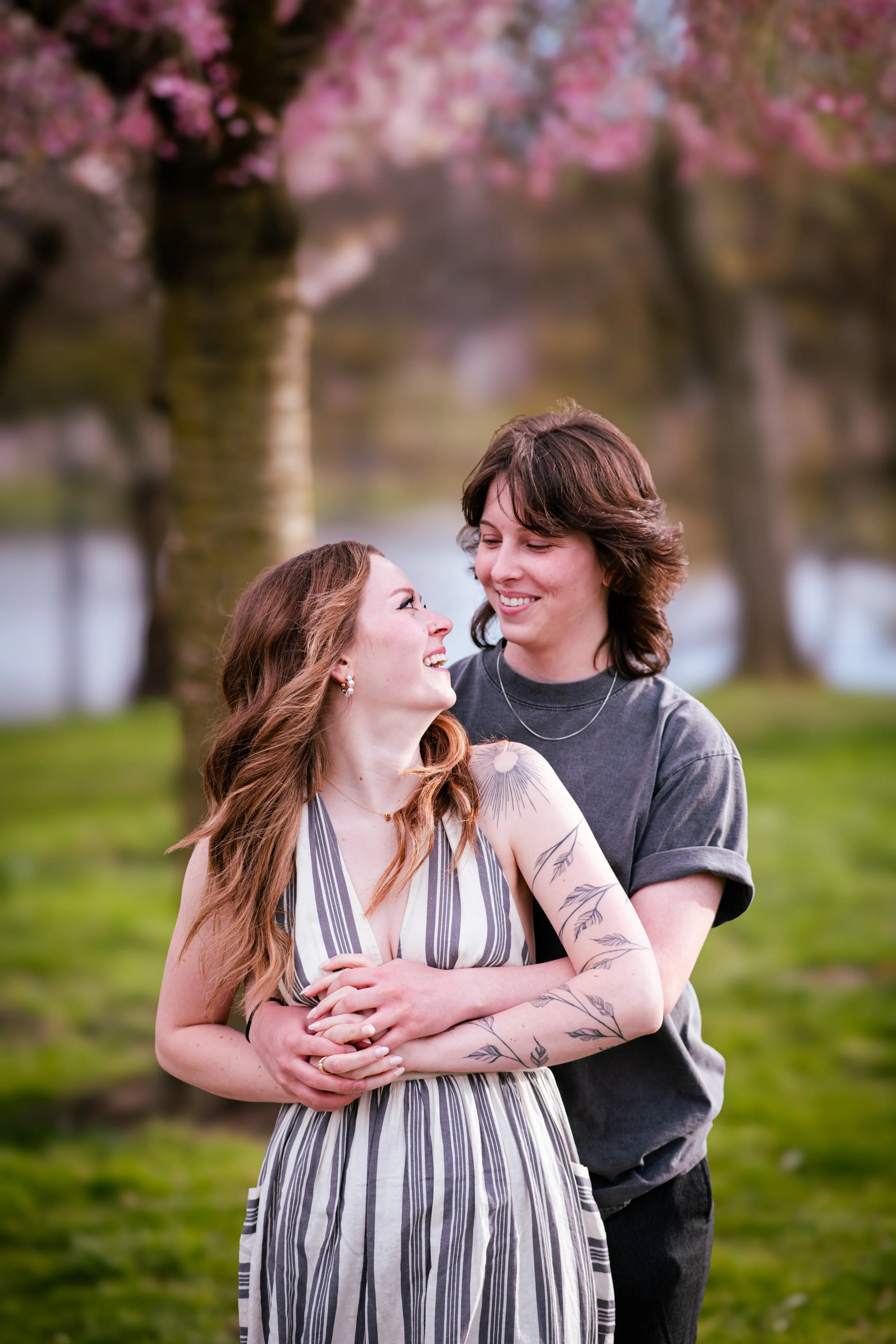 A couple in an outdoor park with blooming pink cherry blossom trees, smiling and embracing each other.