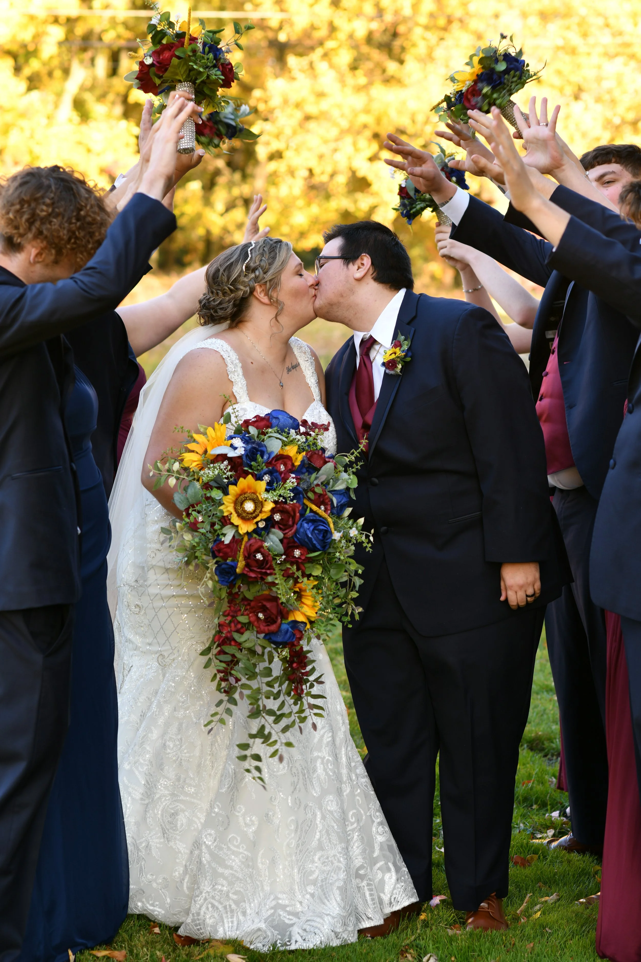 A bride and groom kiss surrounded by wedding guests forming an arch with their hands, outdoors with autumn foliage in background.