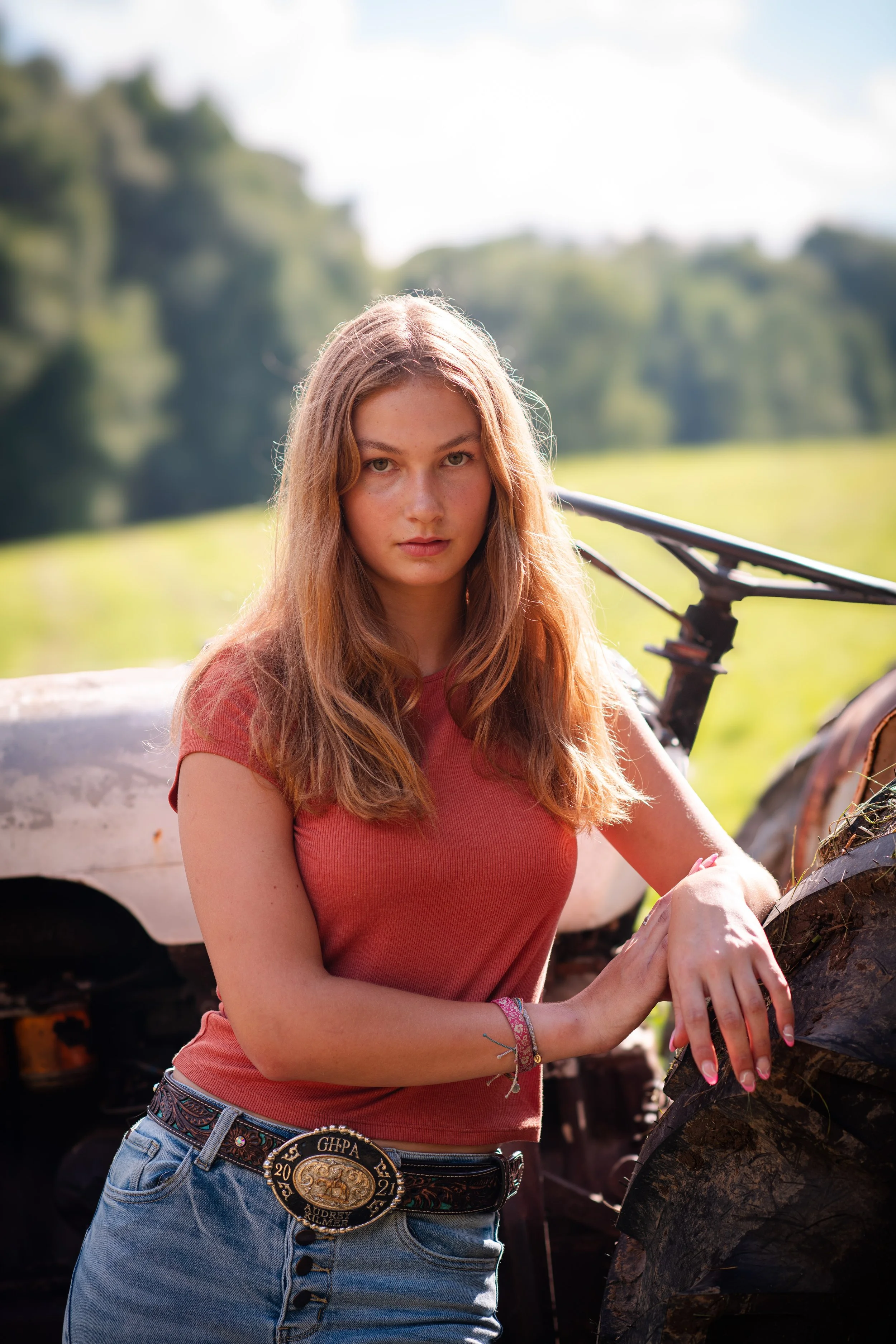 A young woman with long, wavy auburn hair, wearing a rust-colored T-shirt and jeans, standing outdoors with a grassy background and an old, rusty vehicle.