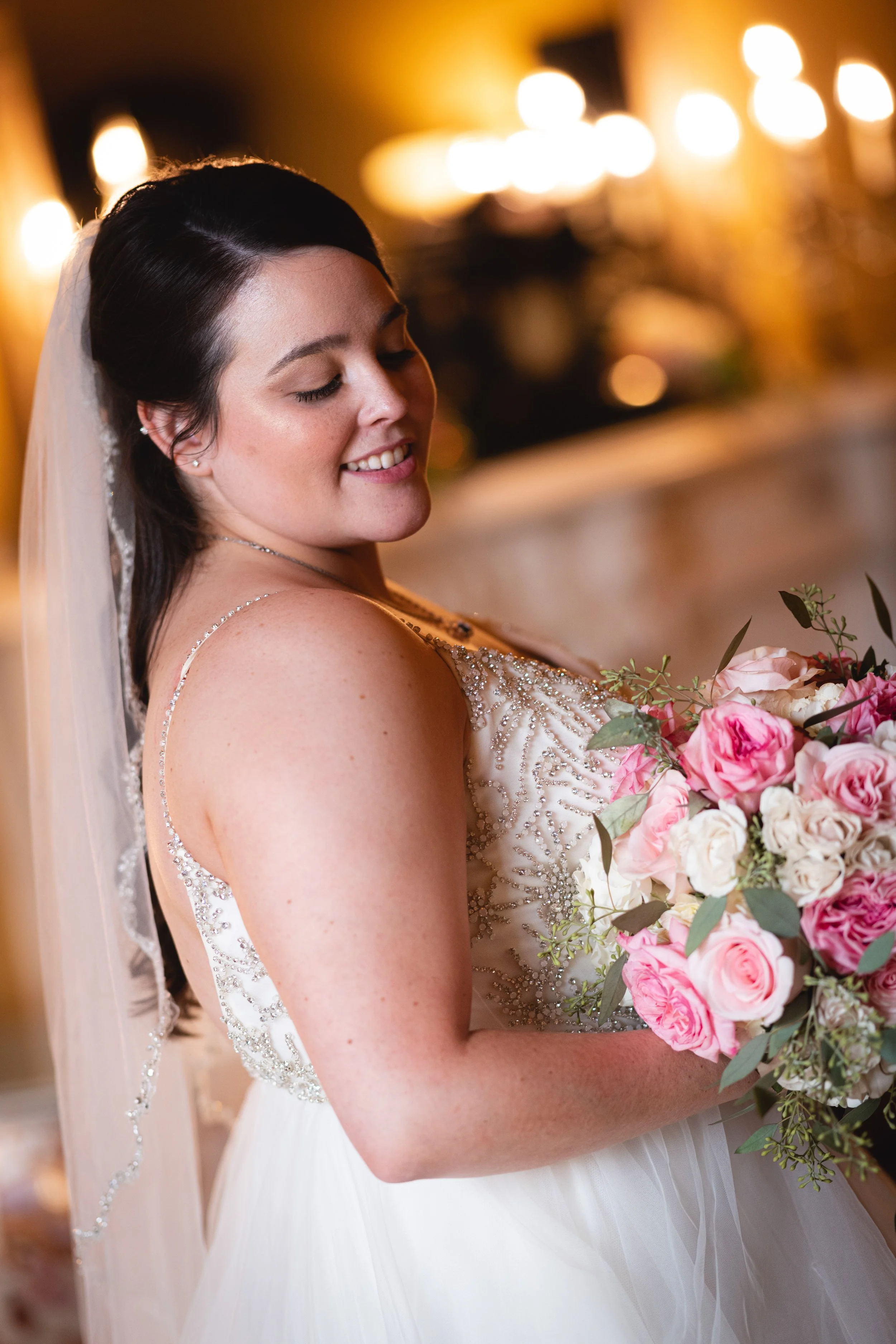 A bride with dark hair, smiling and holding a bouquet of pink and white roses, in a wedding dress with intricate beadwork, indoors under warm lighting.