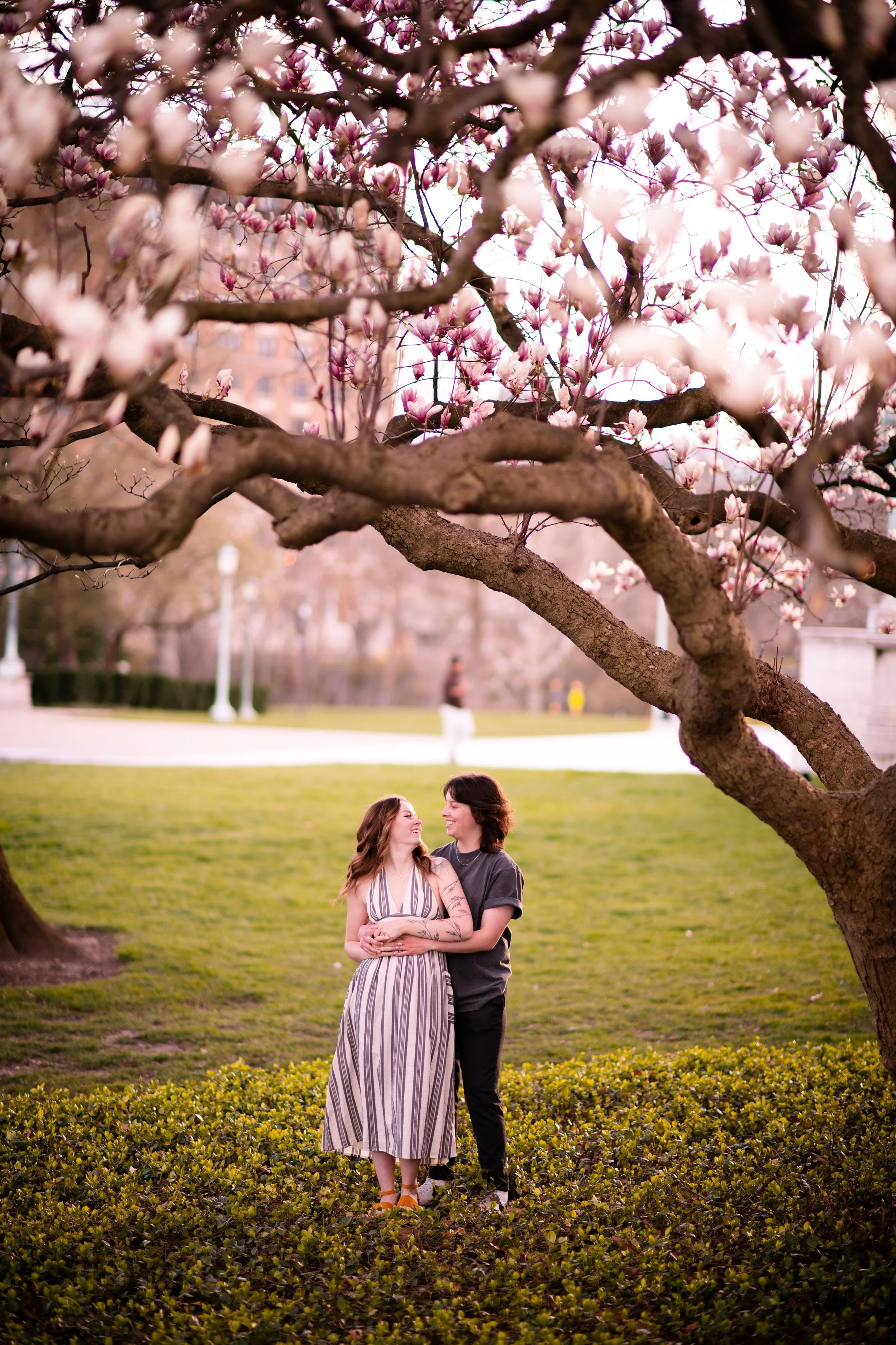Two women standing under a blooming pink magnolia tree in a park, smiling at each other.