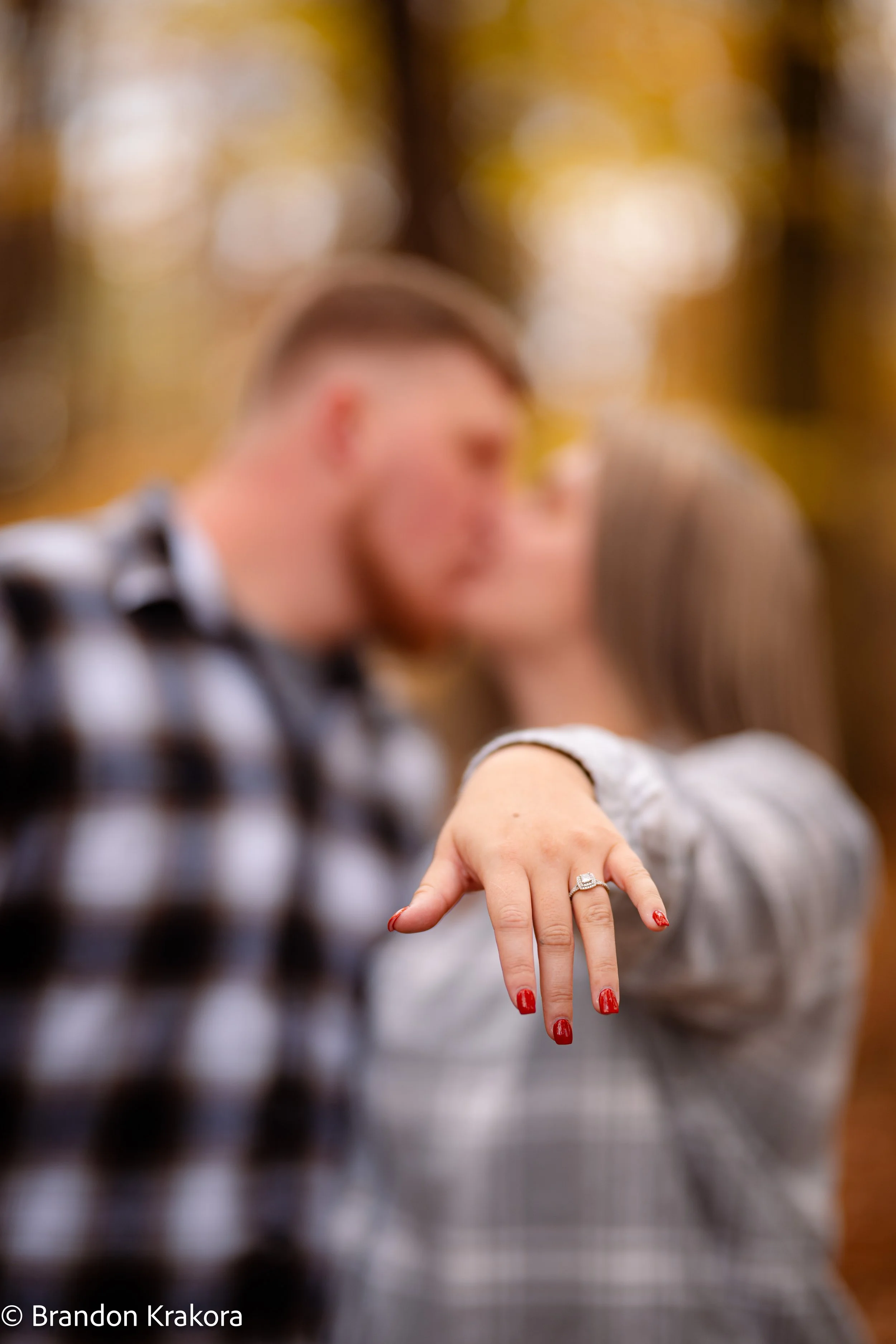 A couple is about to kiss in the background, with a focus on the woman's hand showing an engagement ring and red nail polish.
