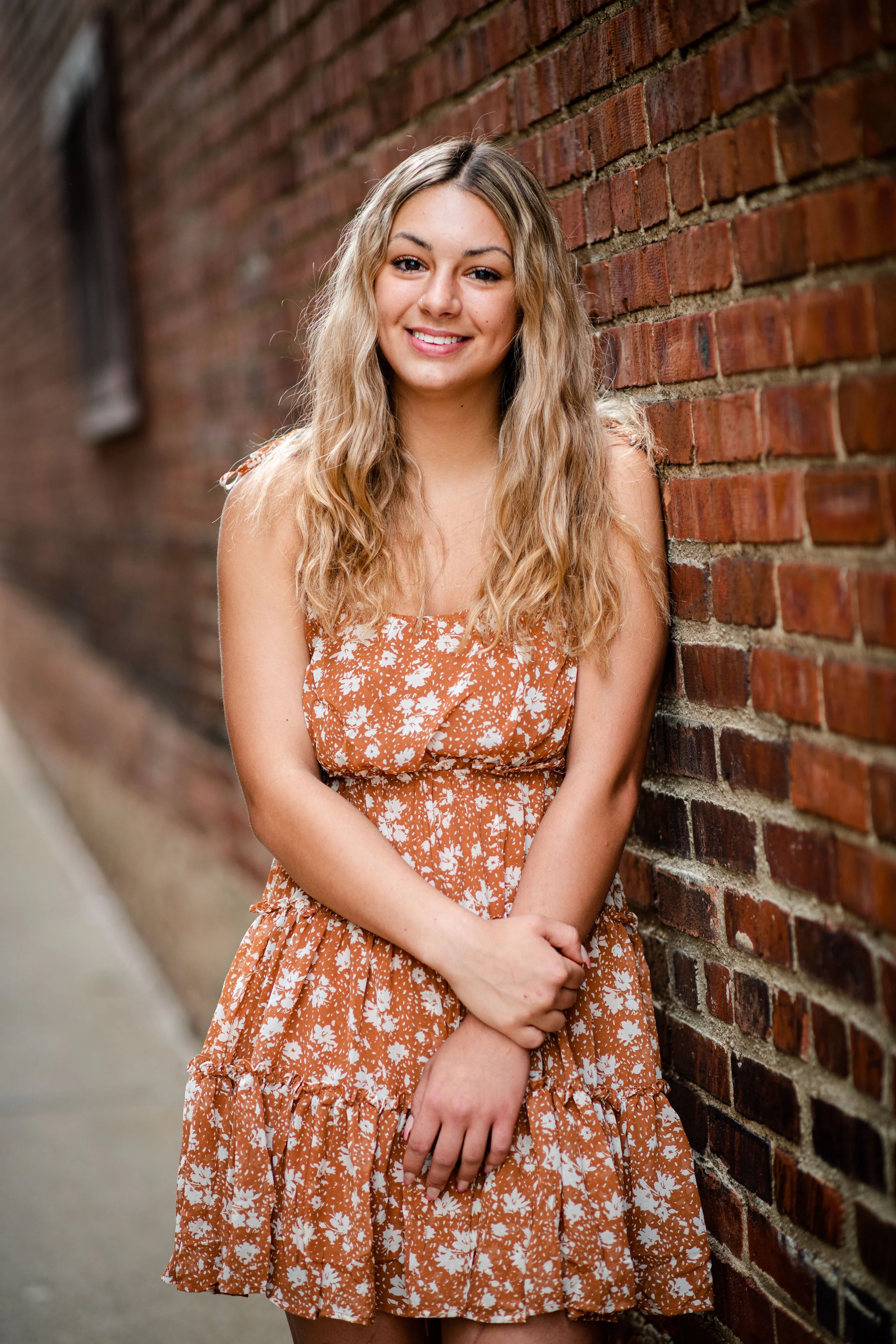A young woman with long, wavy blonde hair smiling, standing next to a brick wall on a city sidewalk.