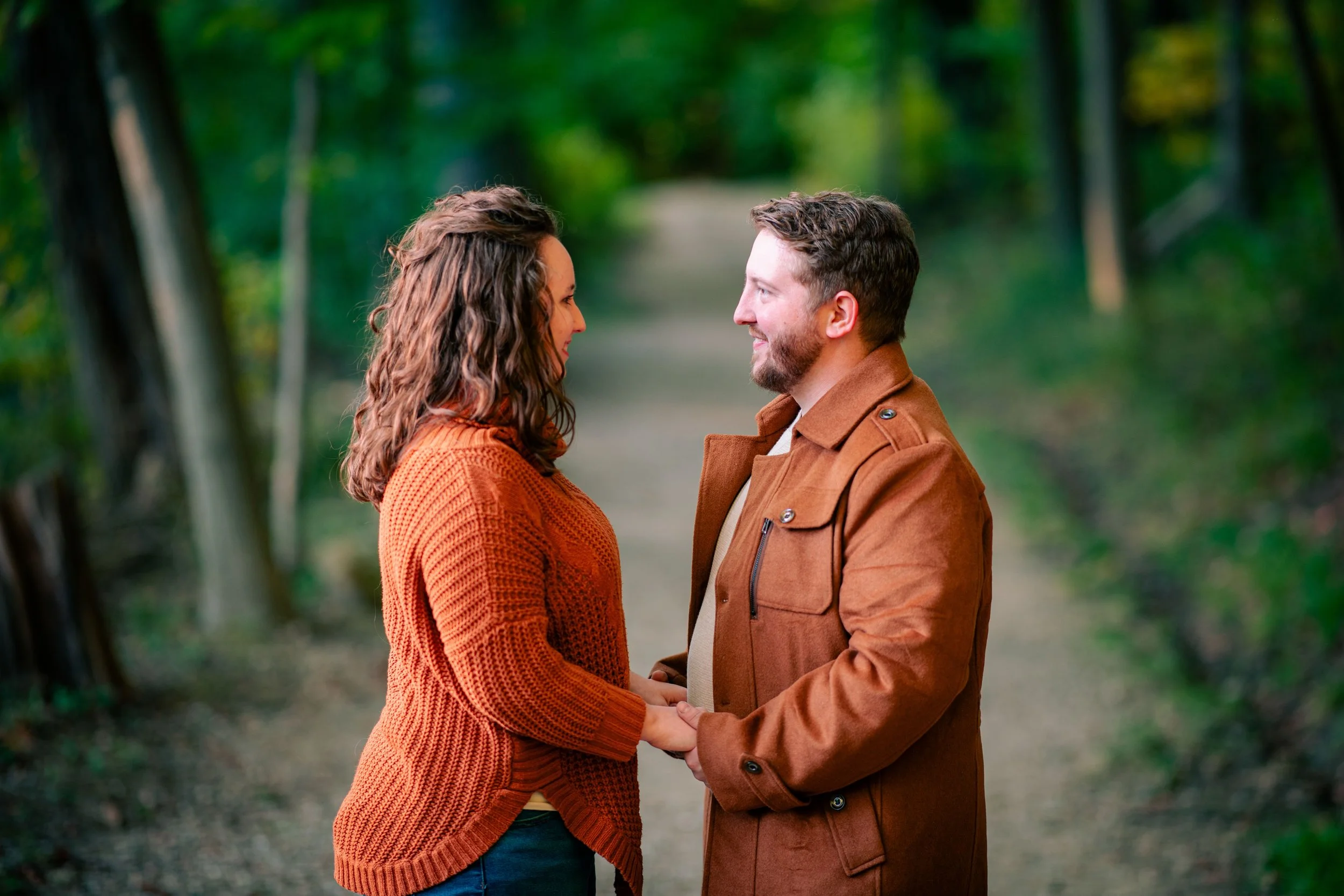 A man and woman standing face-to-face on a forest path, holding hands and looking at each other lovingly.