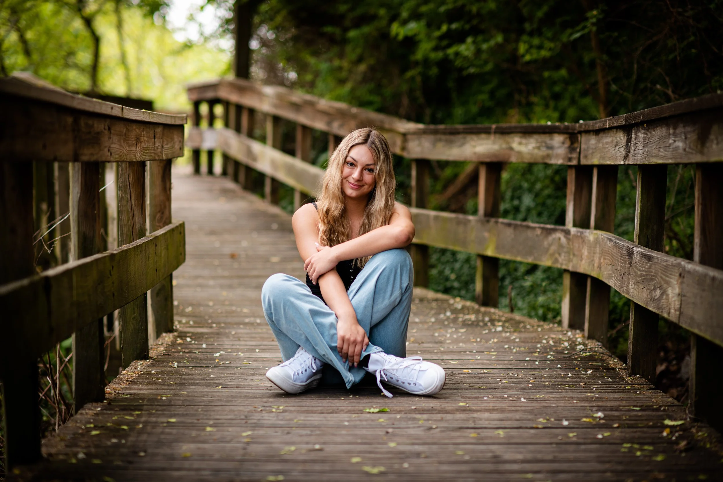 A young woman with long blonde hair sitting on a wooden bridge surrounded by green trees, smiling at the camera.