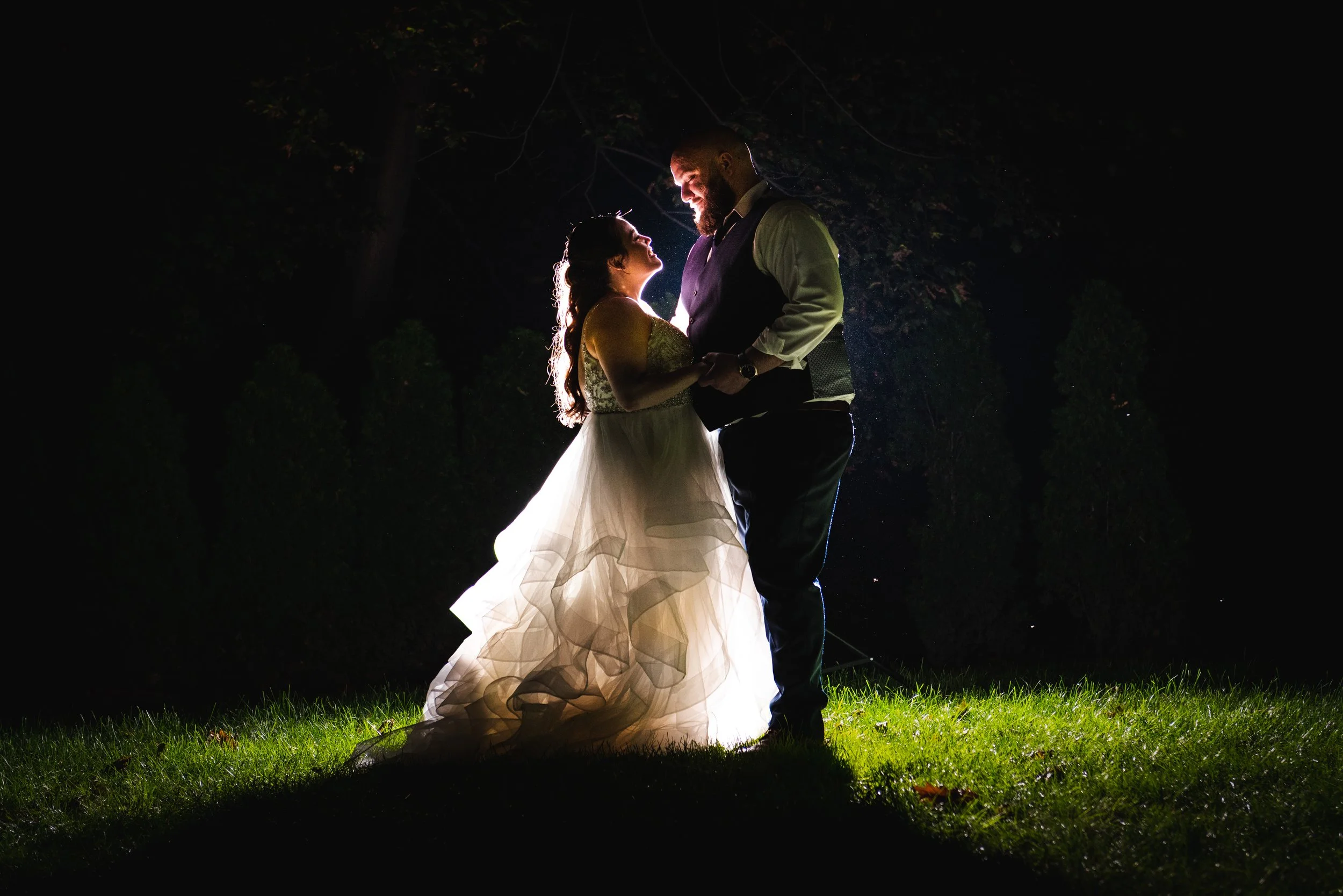 A bride and groom dancing at night outdoors, backlit with a bright light behind them, on a grassy area.