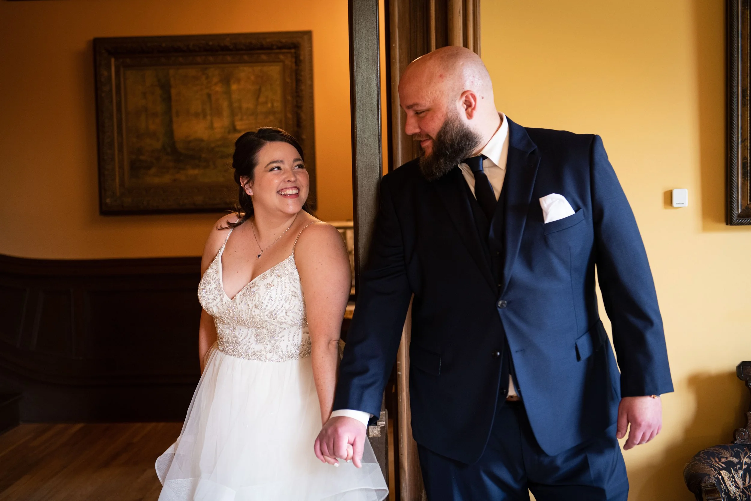 A bride in a white wedding dress and a groom in a dark suit holding hands and smiling at each other indoors.