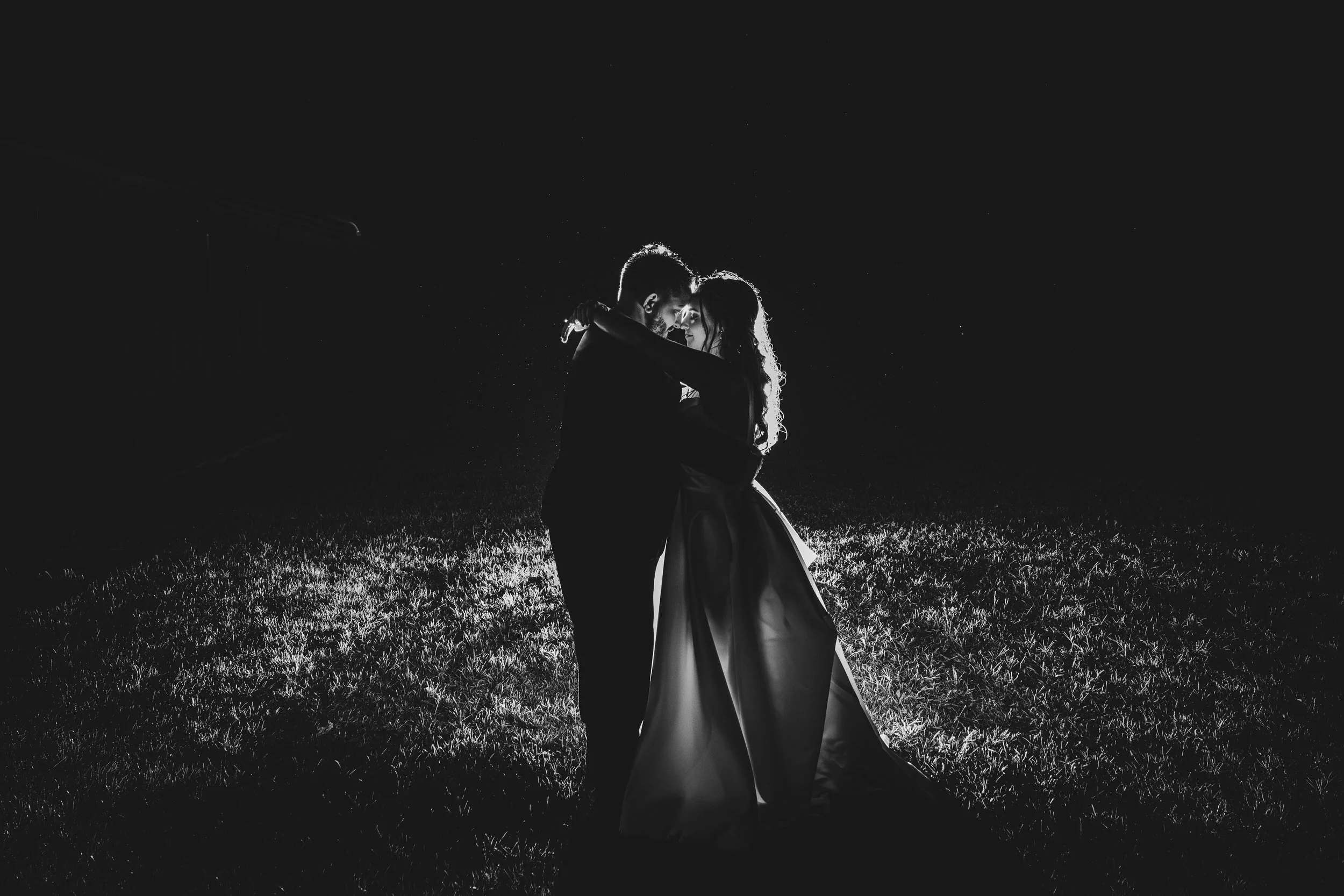 Silhouette of a couple dancing, glowing from behind, on a grassy field at night, stars visible in the sky.