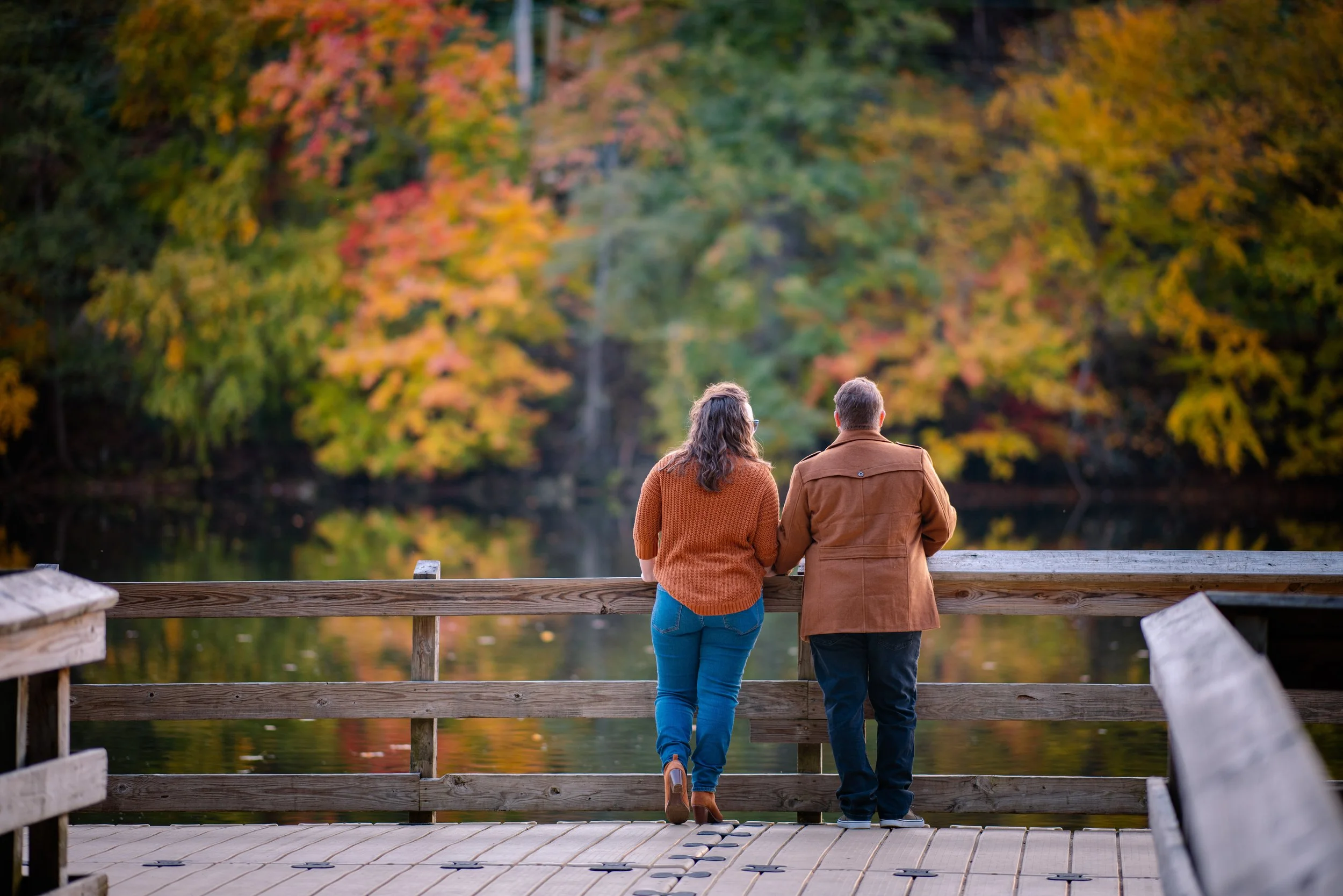 A couple standing on a wooden dock, leaning on a railing, overlooking a lake with colorful fall foliage in the background.