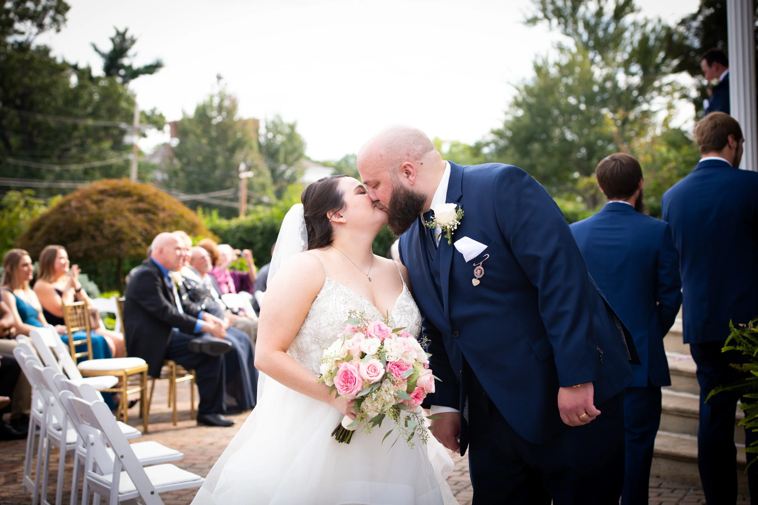 A bride and groom share a kiss at their outdoor wedding ceremony, with guests sitting in chairs behind them.