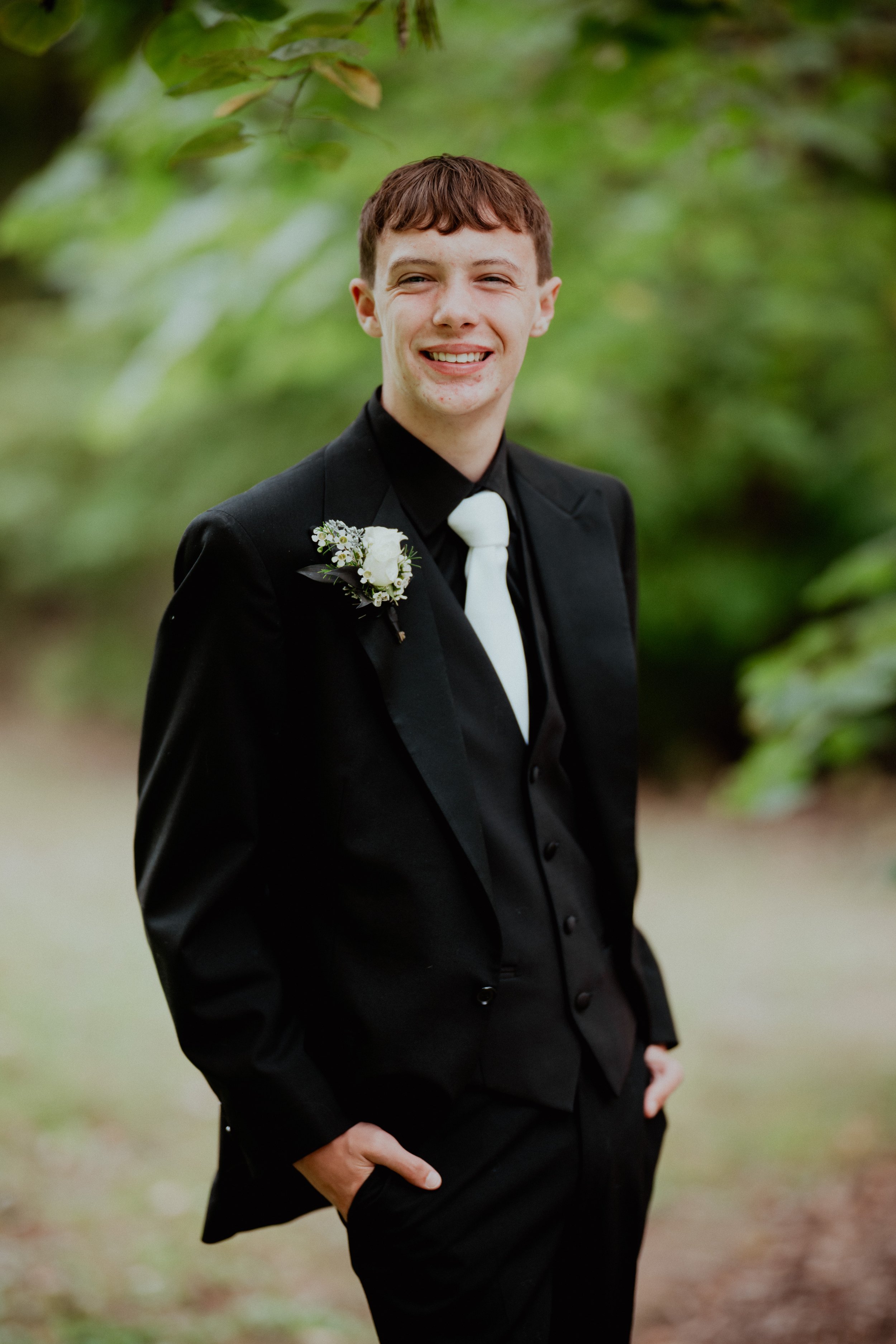 A young man in a black tuxedo with a white tie is standing outdoors in front of greenery, smiling with hands in pockets, and has a boutonniere on his left lapel.