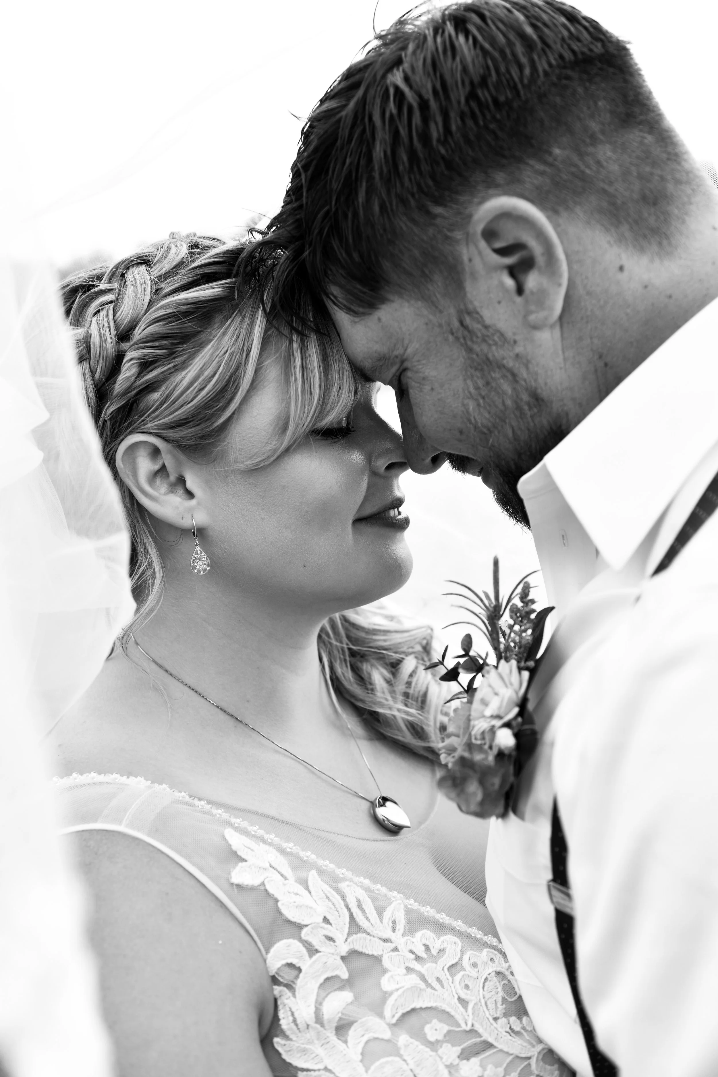 A black and white photo of a couple with their foreheads touching, smiling gently at each other, dressed in wedding attire.