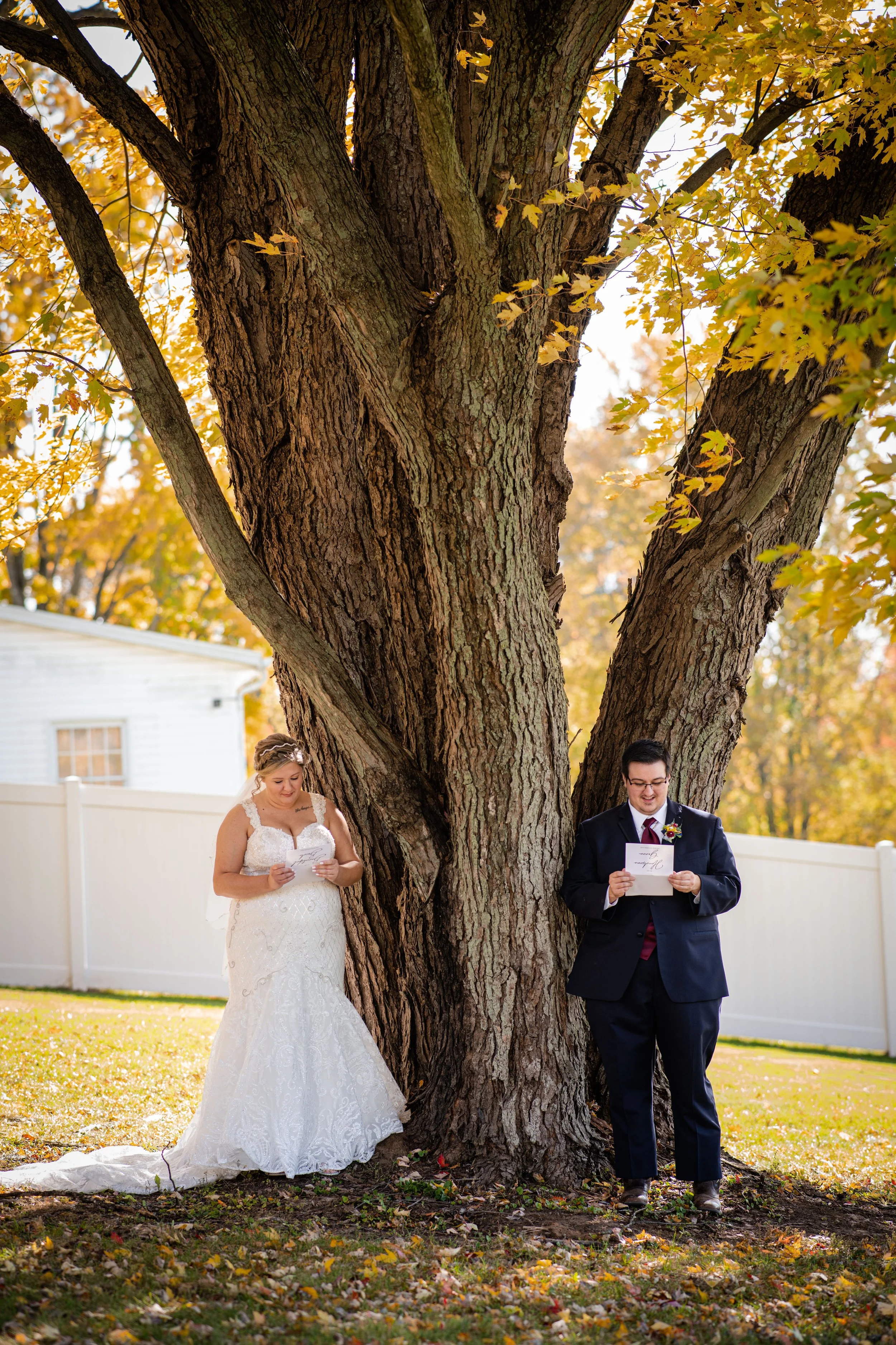 A bride and groom in wedding attire reading vows under a large tree with autumn leaves, outdoor setting with white fence in background.