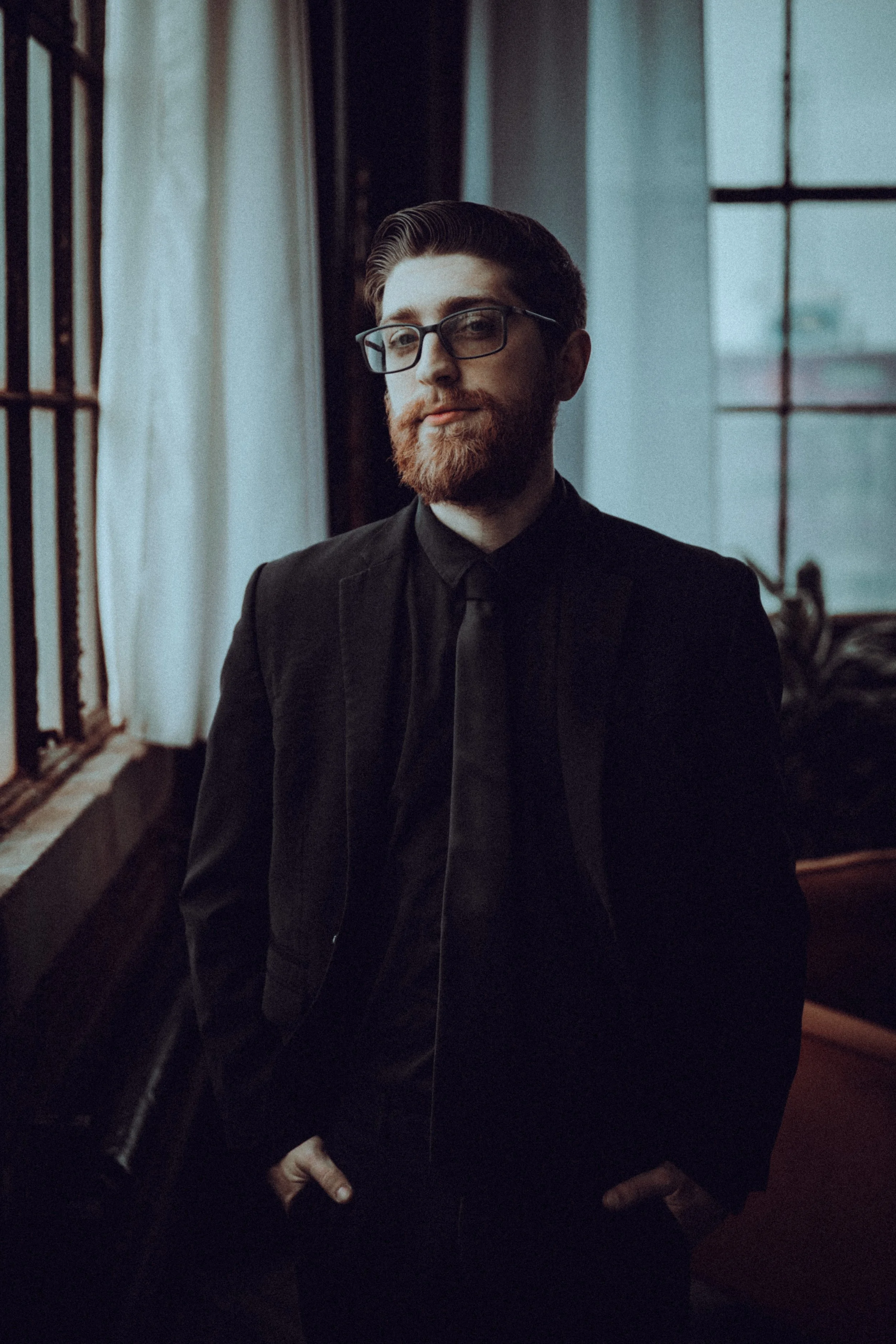 A man with glasses, a beard, and red hair, wearing a black suit and tie, standing indoors near a window with curtains and looking at the camera.