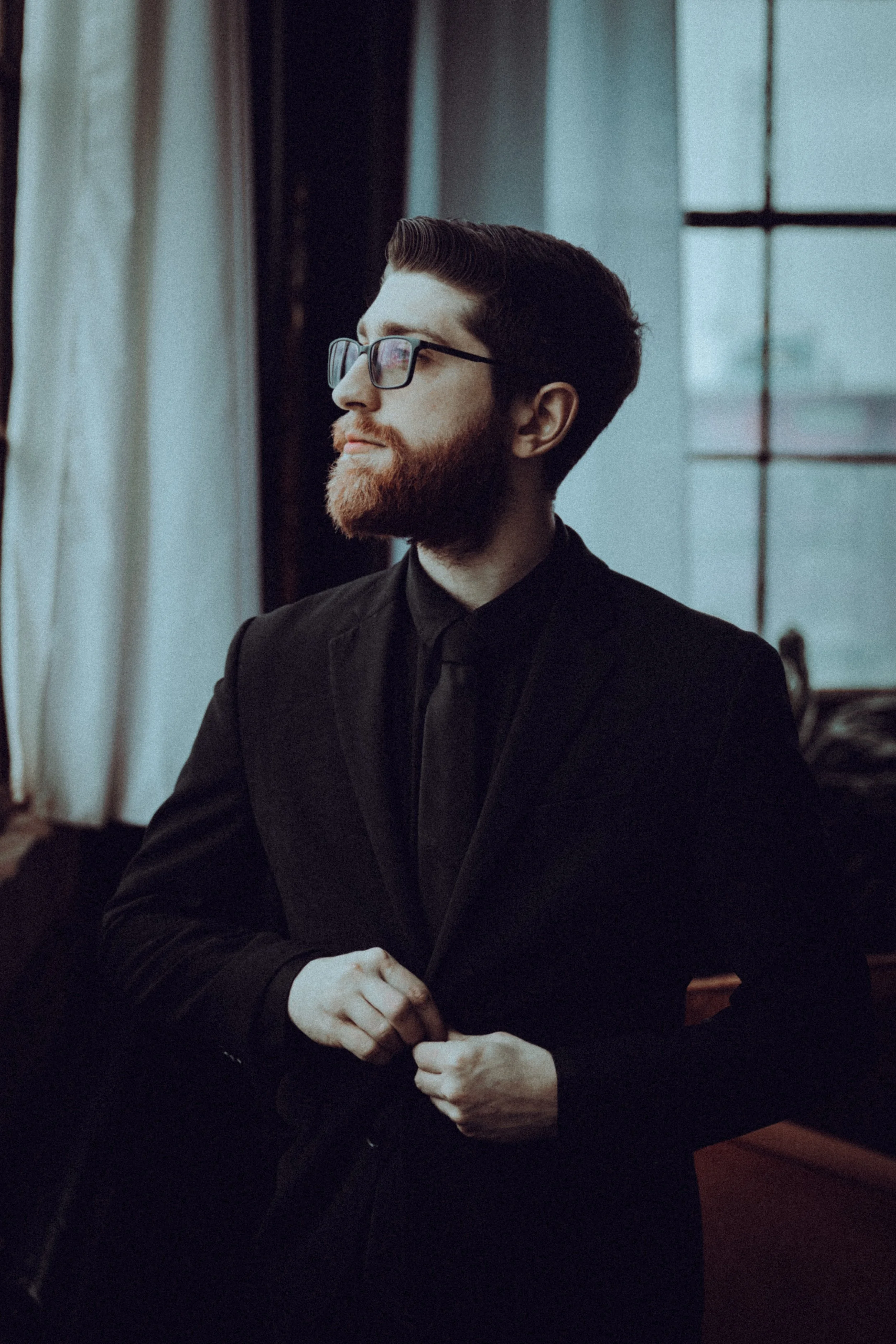 A man dressed in black adjusting his suit jacket, wearing glasses, with a beard, and looking to the side near a window.