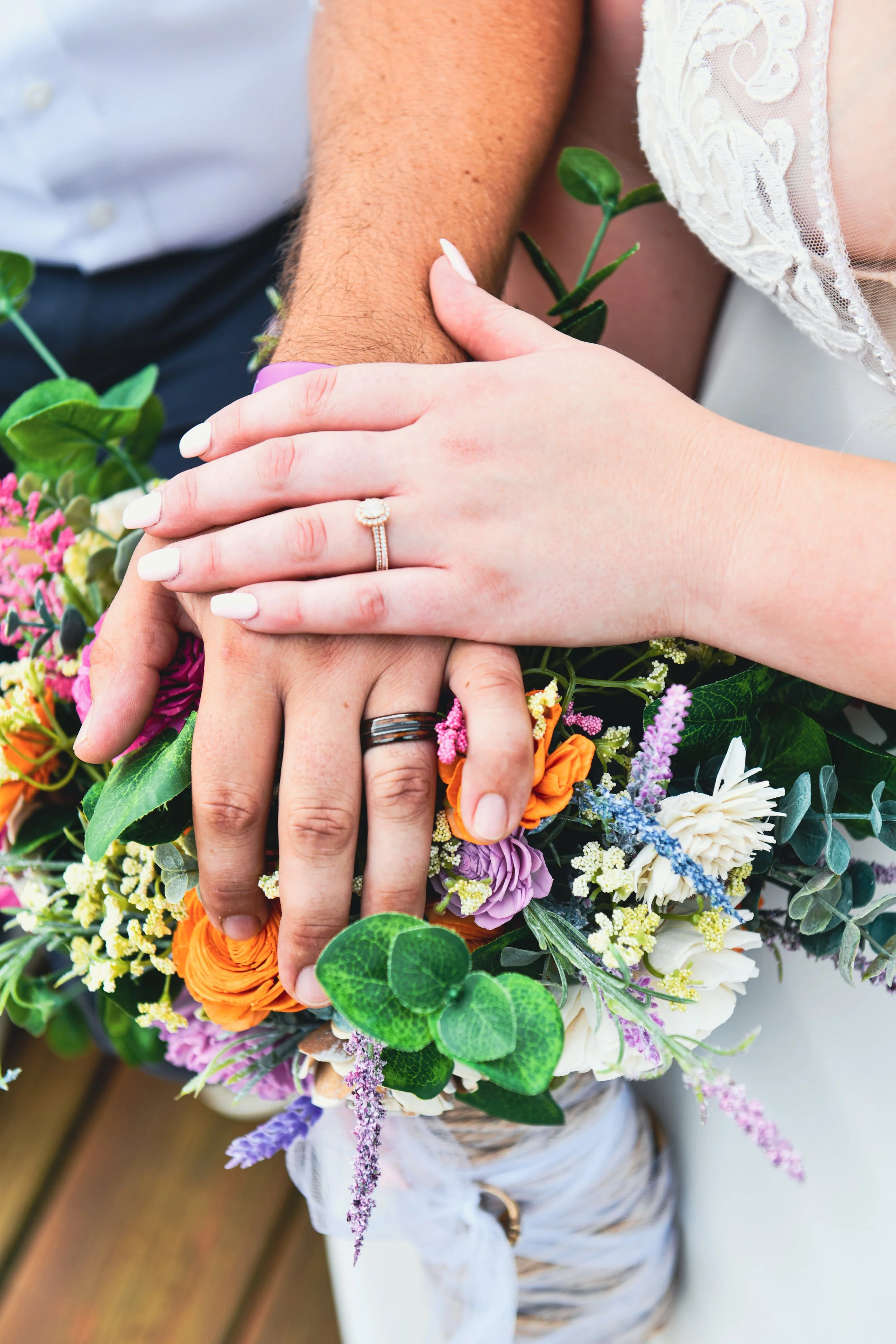 Close-up of a couple's hands with wedding rings, resting on a colorful bouquet of flowers during a wedding celebration.