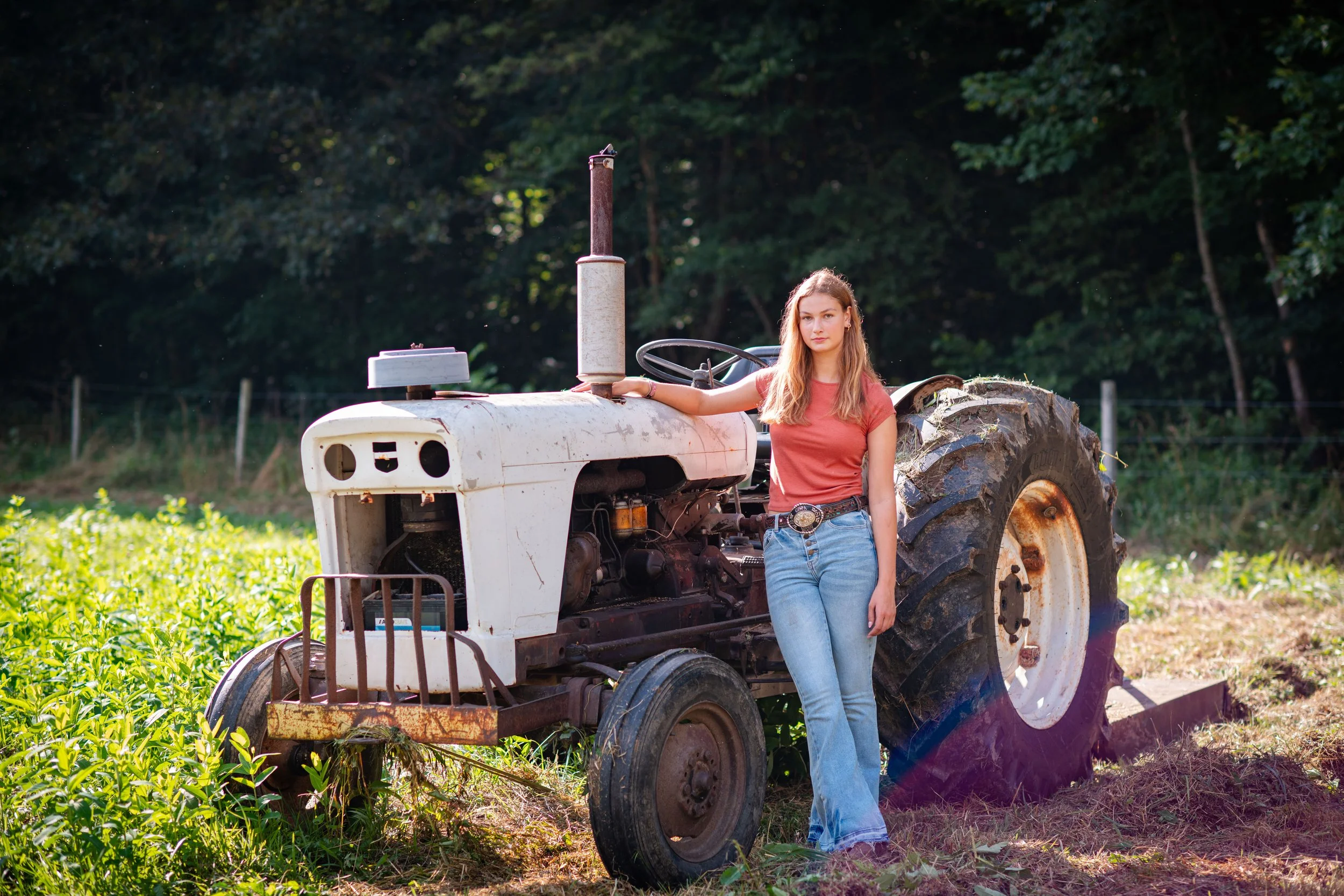 A young woman standing next to an old white tractor in a field with green plants, trees in the background, and sunlight shining on her and the tractor.