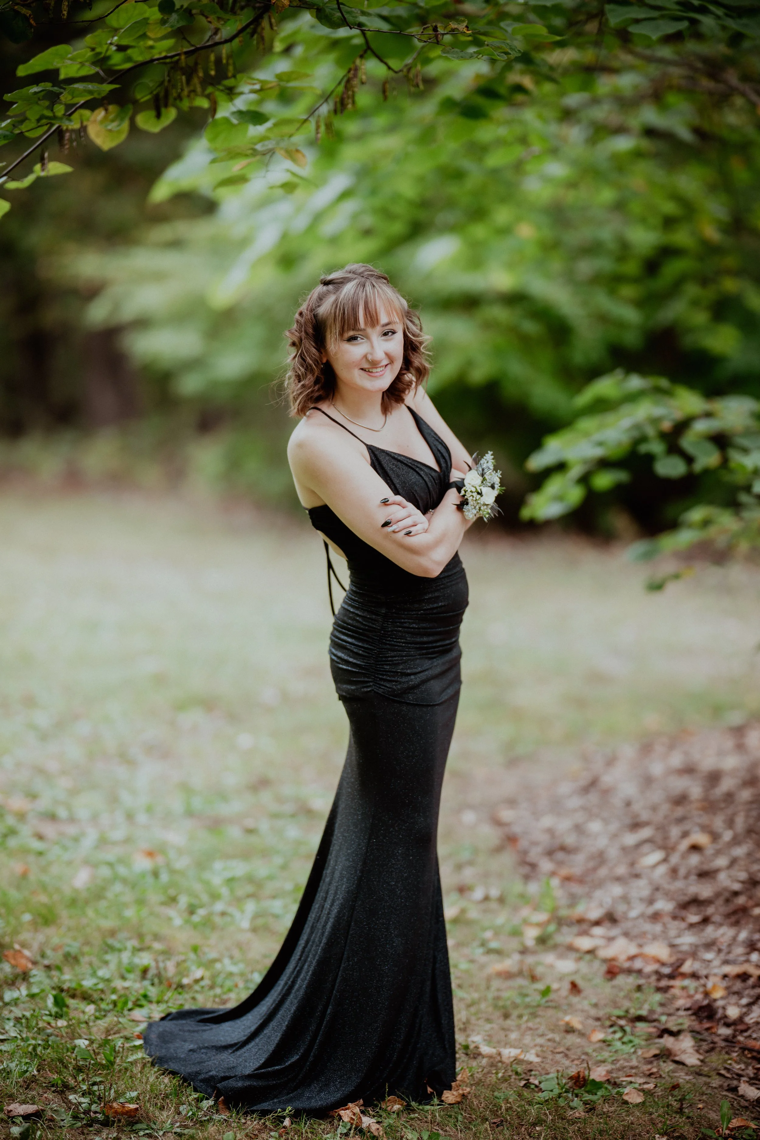 A young woman in a long, black dress standing outdoors among green foliage, smiling with arms crossed, holding a small bouquet of flowers.