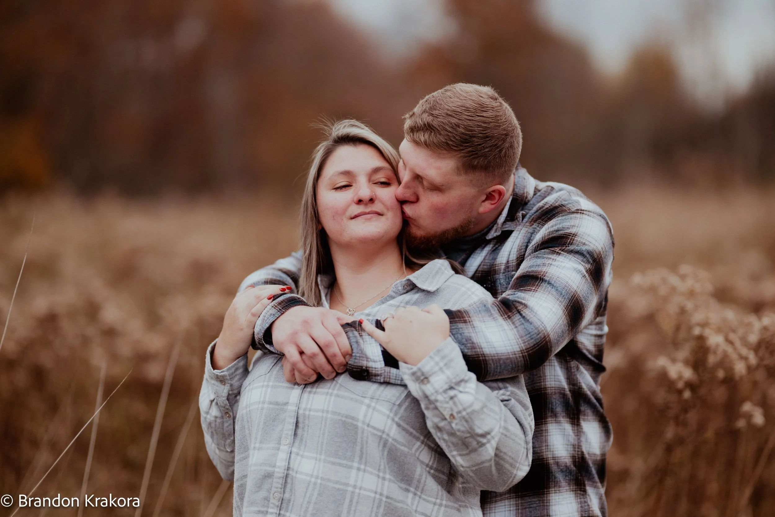 A young couple sharing a tender kiss outdoors in a field during fall, with the man hugging the woman from behind.