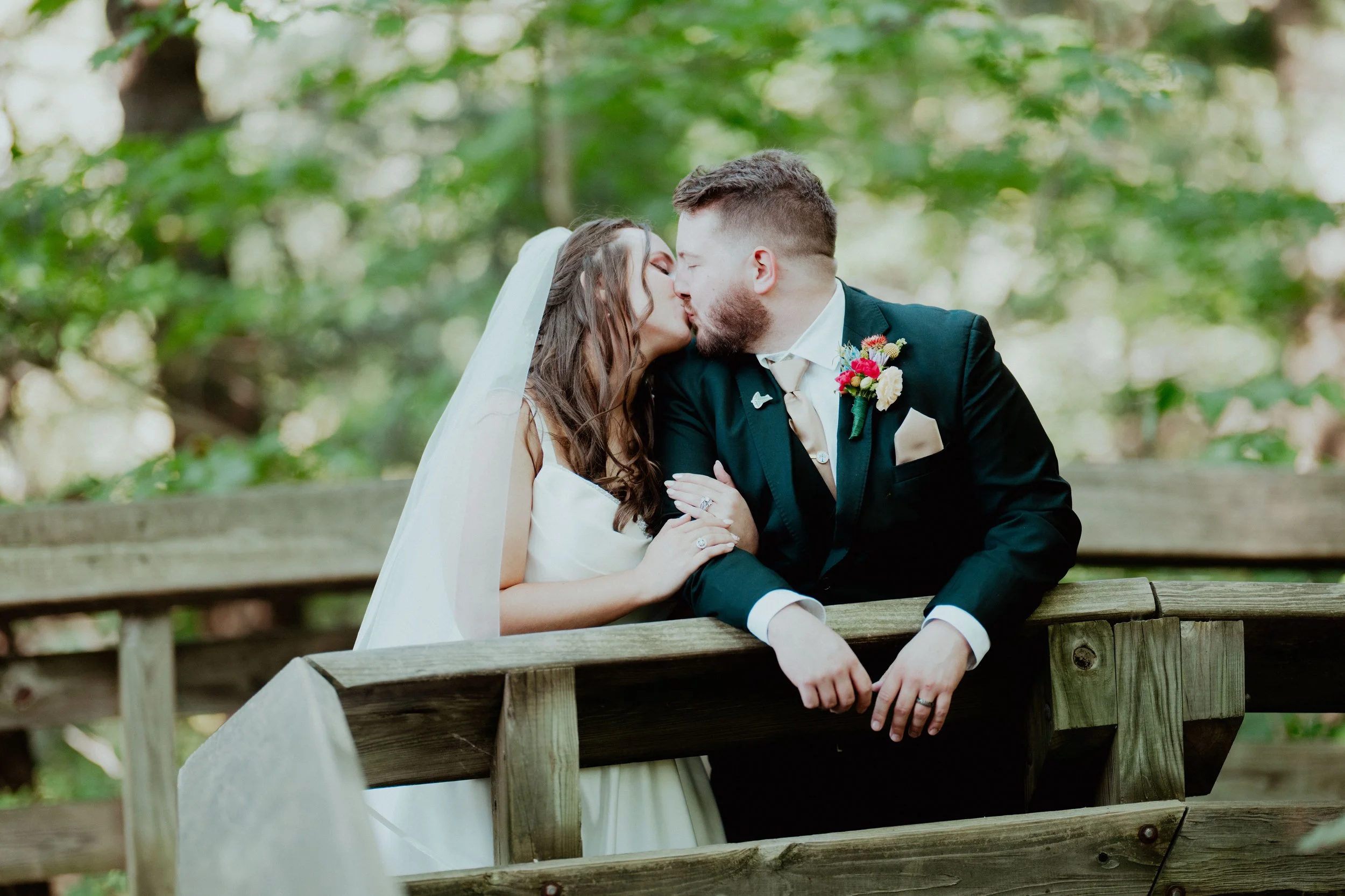 Bride and groom sharing a kiss on a wooden bridge outdoors with green trees in the background.