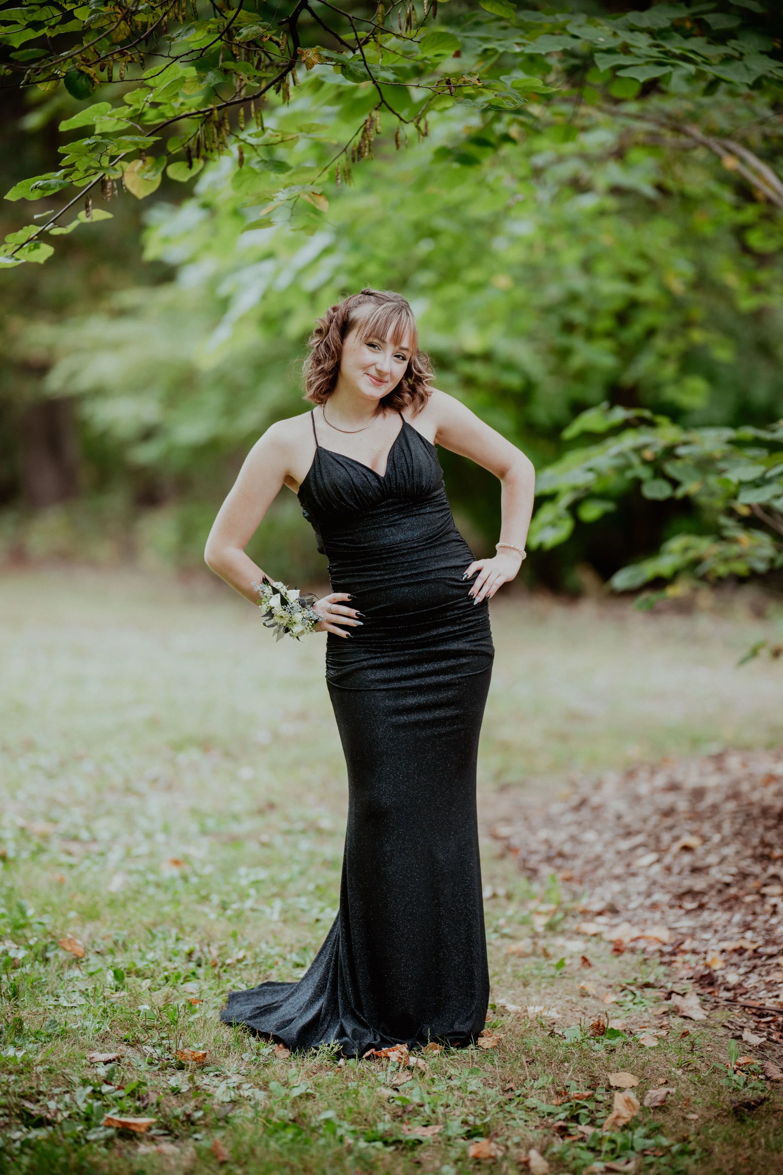 A young woman in a long, form-fitting black dress with spaghetti straps standing outdoors in a wooded area with green trees and grass, smiling with her hands on her hips.