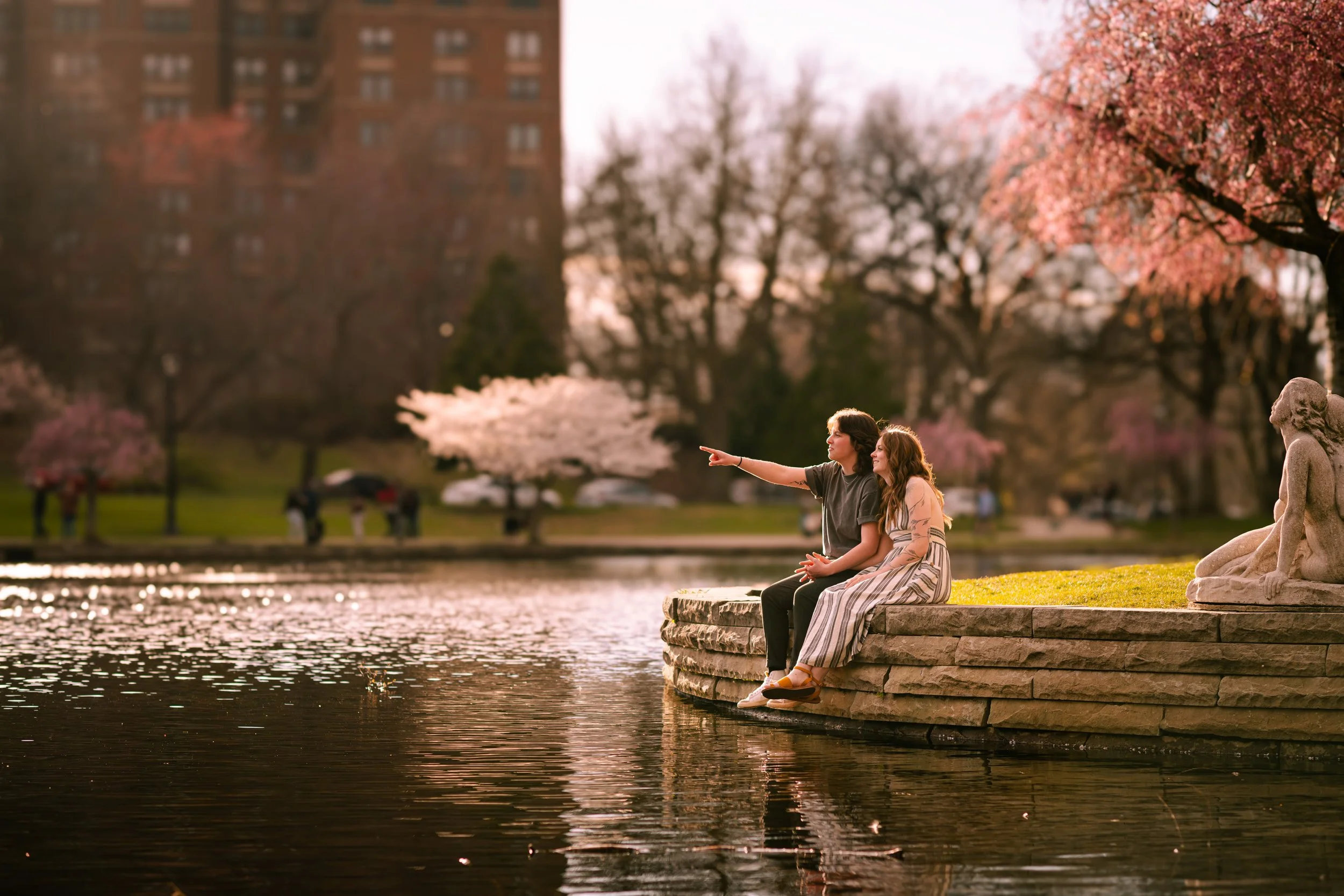 A boy and girl sitting on a stone edge by a pond, surrounded by blooming cherry blossom trees in a park at sunset.