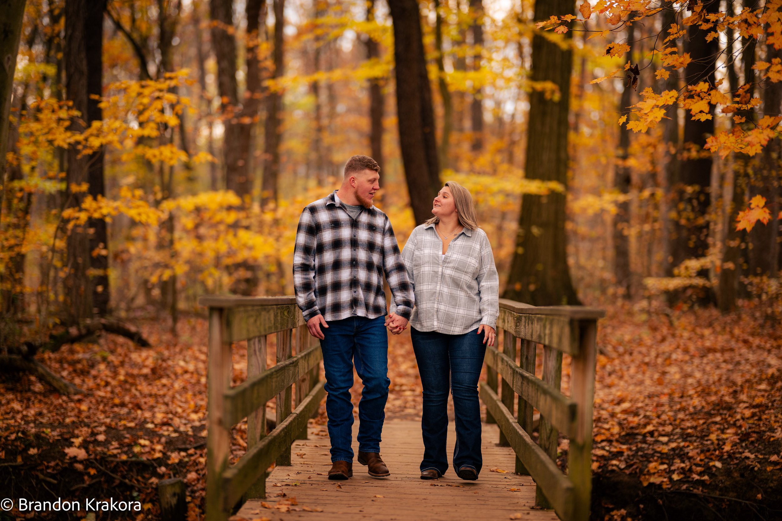 A couple holding hands and walking on a wooden bridge through a forest with autumn leaves.