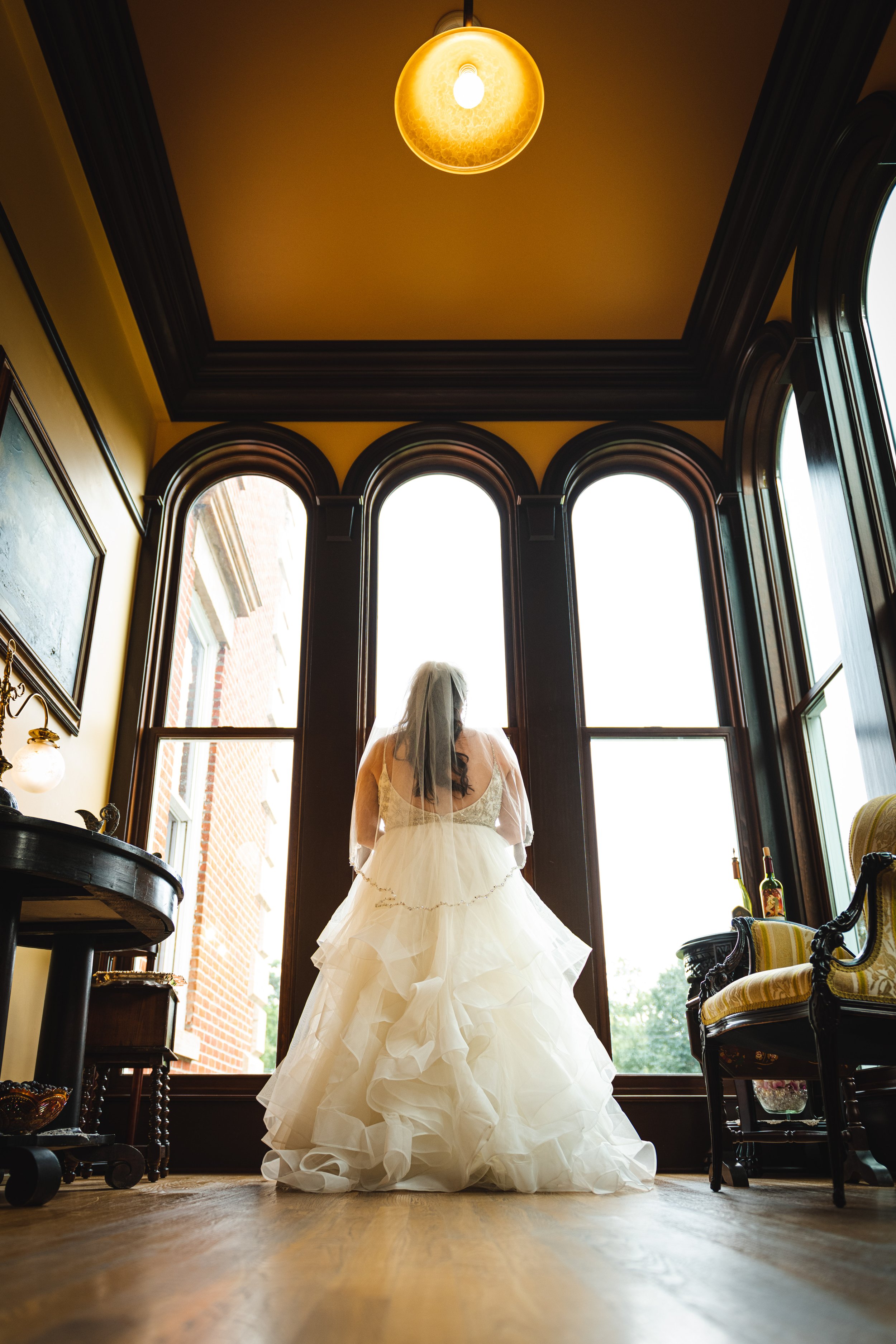 Bride in a wedding dress and veil standing indoors near large arched windows, illuminated by a ceiling light, with antique furniture and bottles in the background.