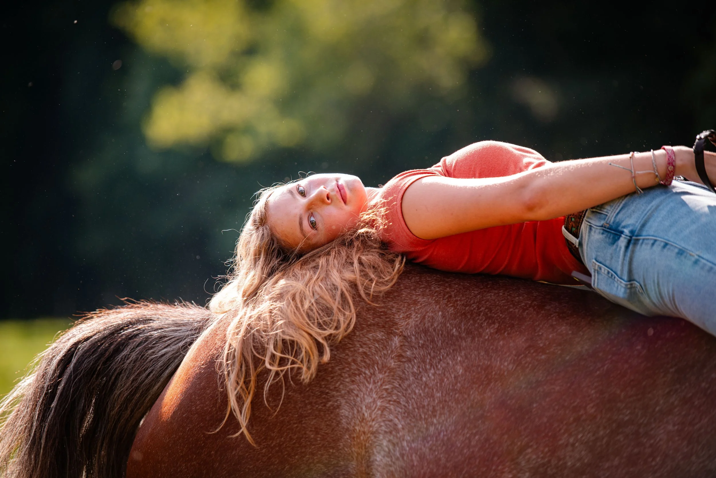 Young woman lying on a brown horse outdoors, looking at the camera with sunlight and greenery in the background.