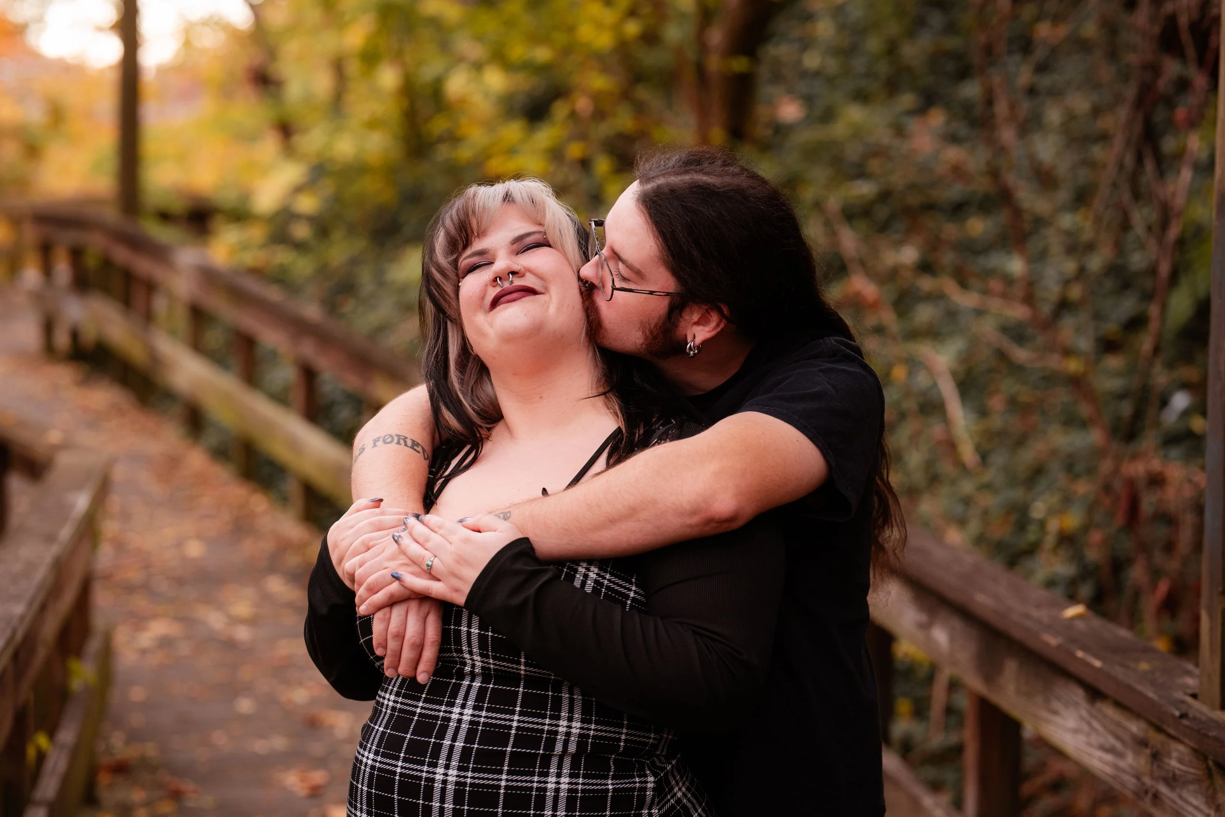 A man kissing a woman on the cheek while hugging her from behind on a wooden bridge in an autumn forest.