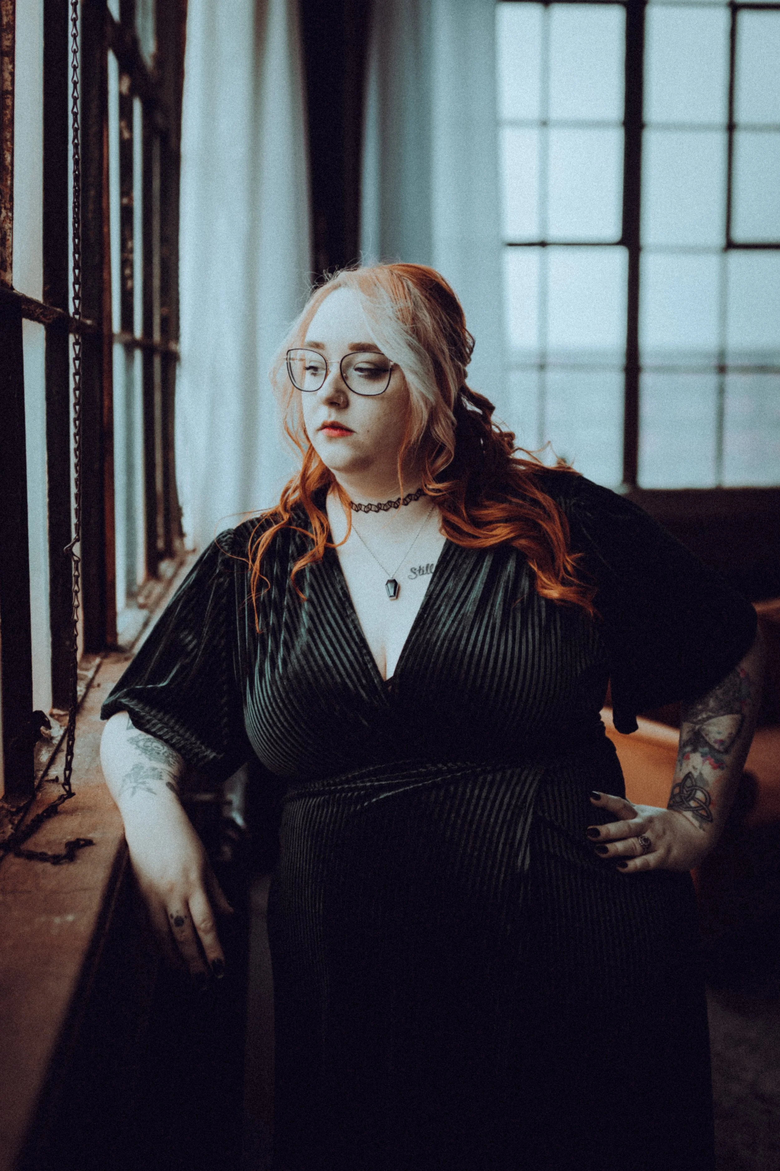 A woman with long red wavy hair and glasses standing near a large window with black metal framing in an industrial-style room. She is wearing a black dress with a V-neck, choker necklace, and tattoos on her arms. She is looking to her left with her h
