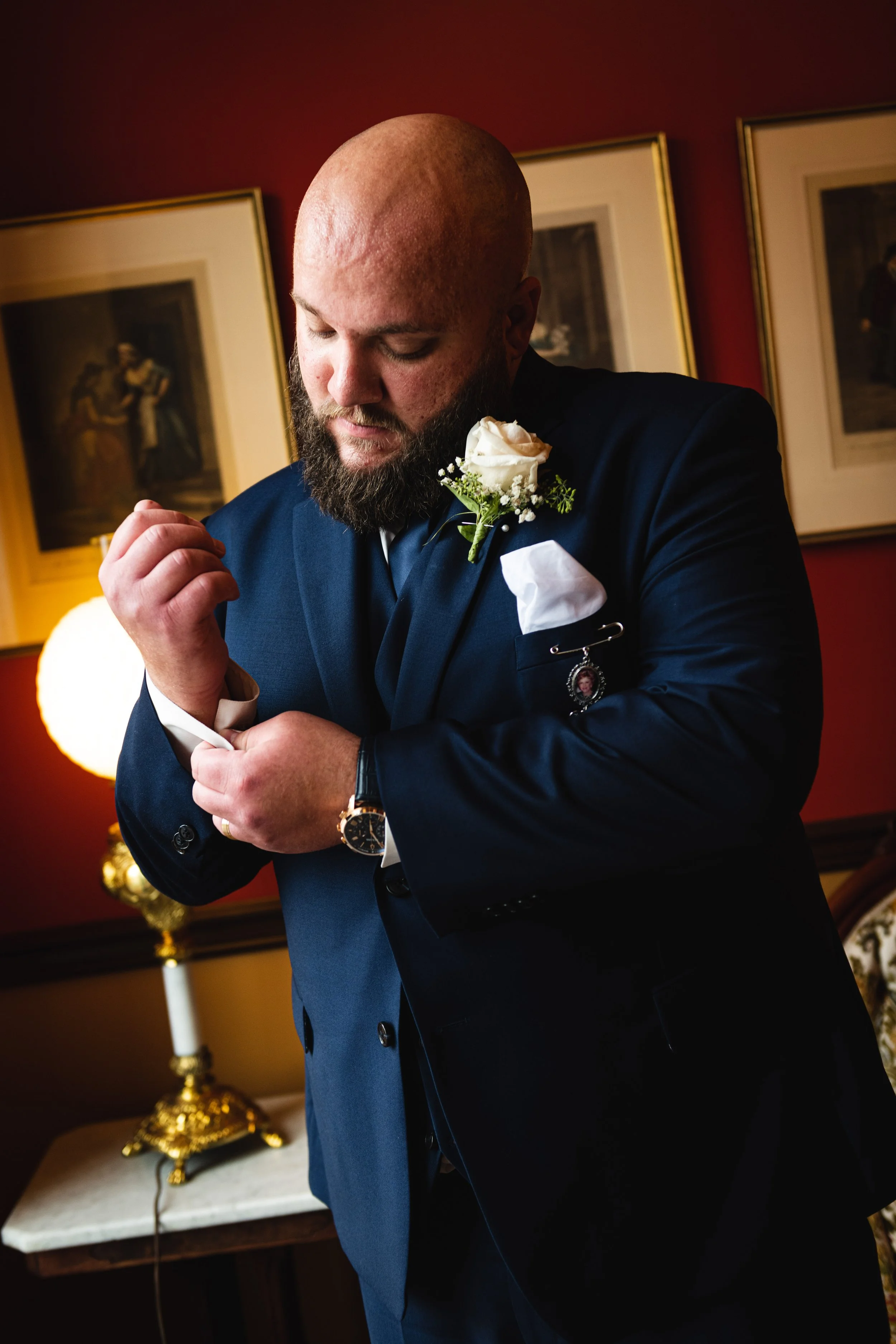 A man with a beard wearing a dark blue suit and white shirt, adjusting his cufflink. He has a white pocket square, a boutonniere with a white rose, and a charm pin on his lapel. The background has framed art on a red wall and a gold-colored lamp.