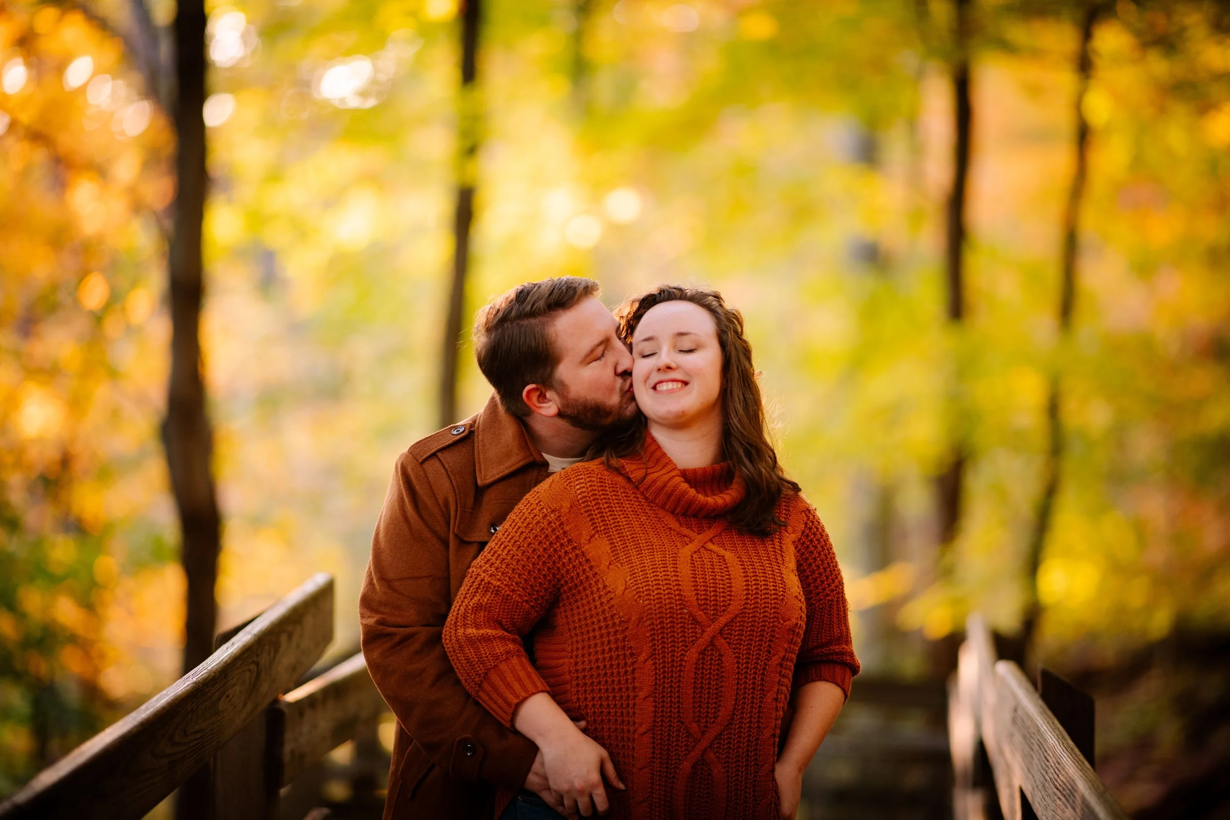A couple embracing on a wooden bridge with autumn foliage in the background. The man kisses the woman on the cheek, and she is smiling with her eyes closed. The woman is wearing an orange sweater, and the man is wearing a brown jacket.