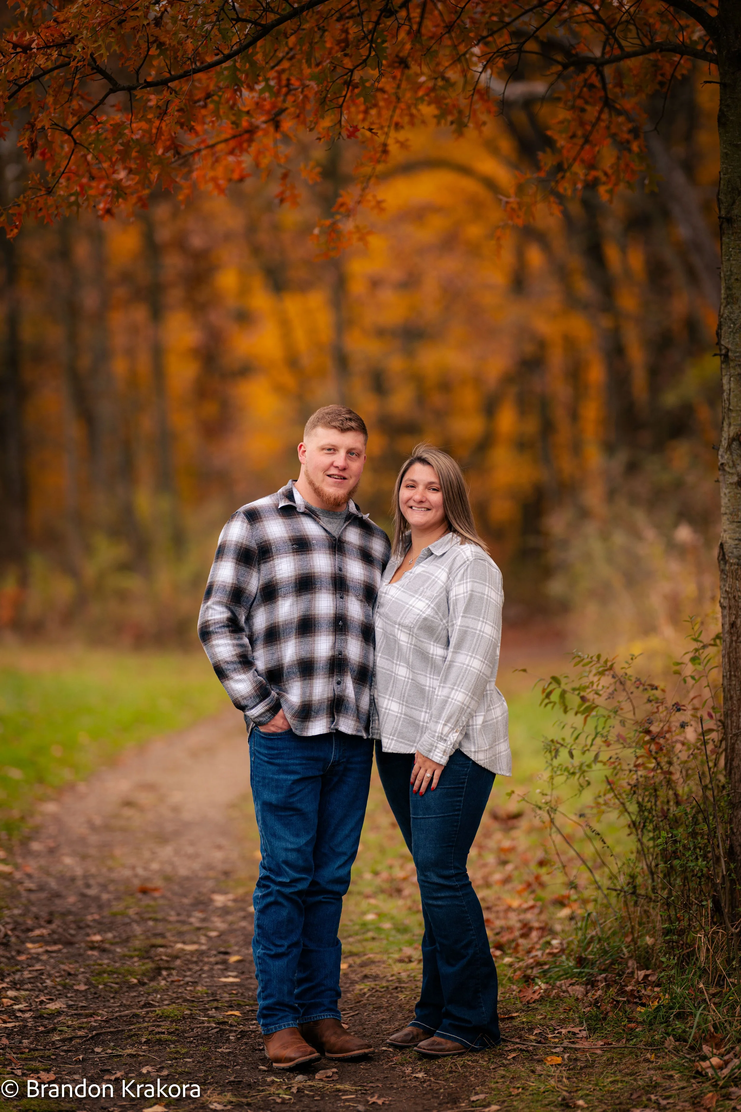 A couple standing on a forest trail during autumn, surrounded by trees with orange and yellow leaves, smiling at the camera.