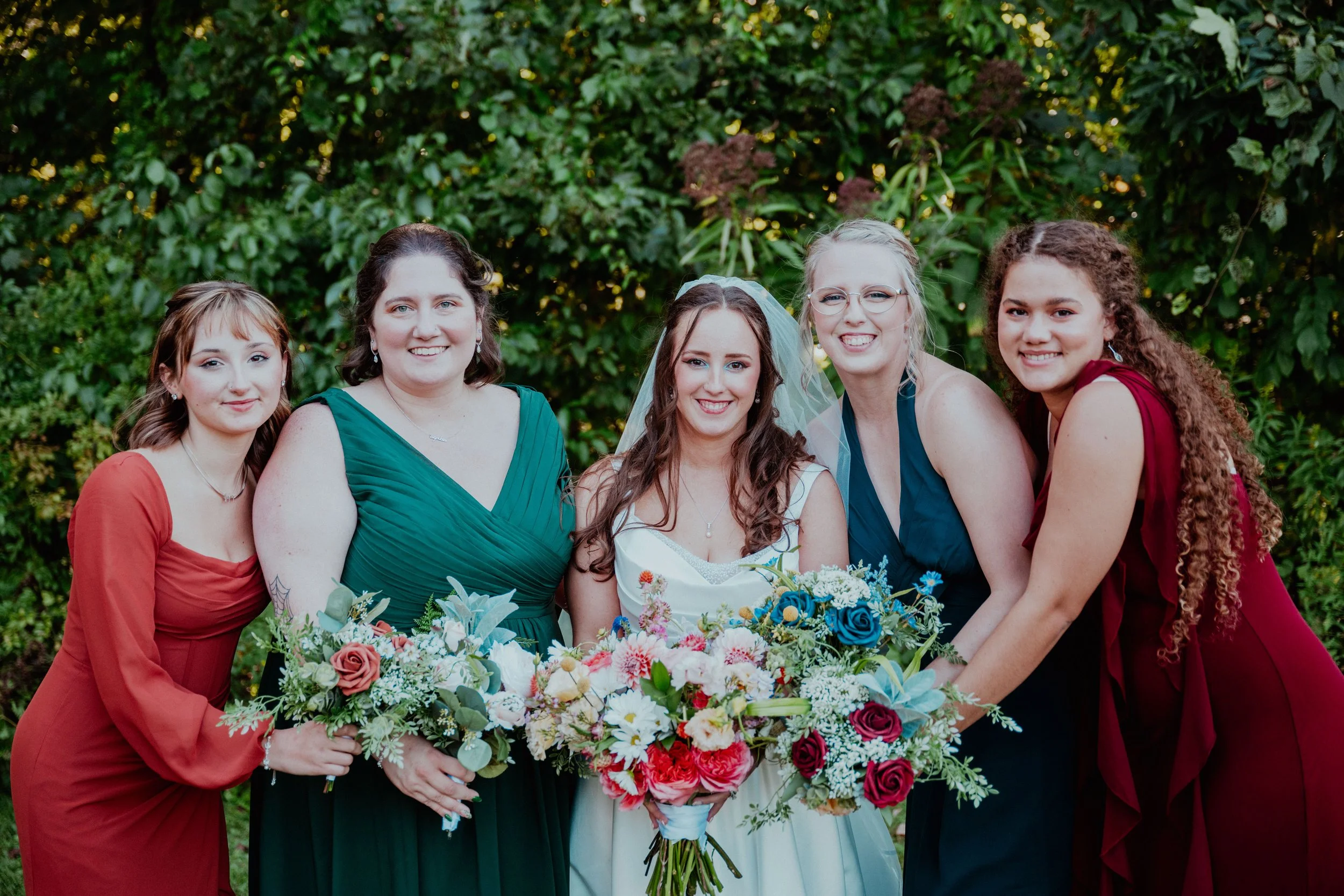 A group of five women at a wedding, standing outdoors with lush green foliage background. The bride in the center is wearing a white wedding dress and veil, holding a large bouquet of colorful flowers. The other four women, likely bridesmaids, are dr