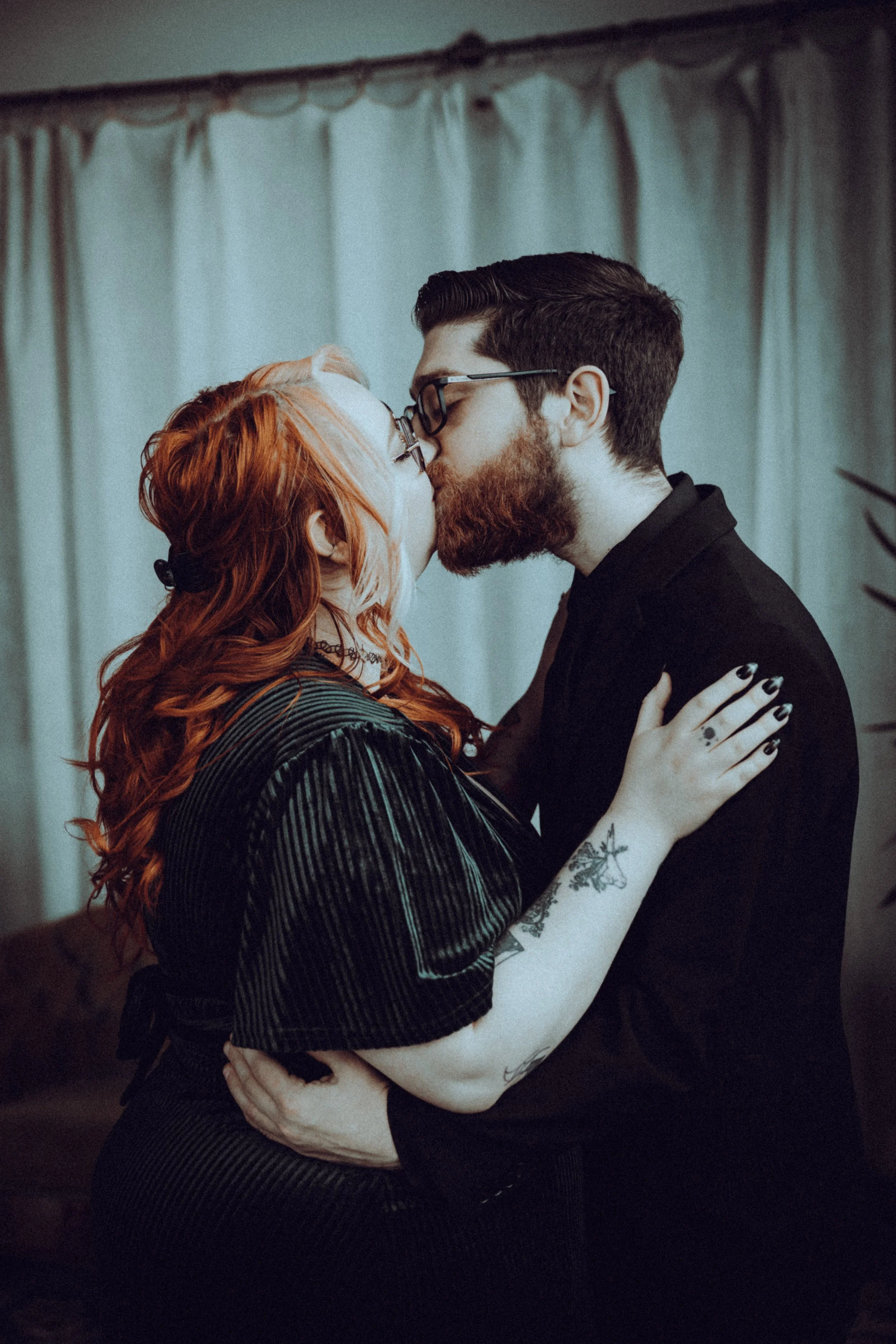 A couple with red hair and glasses kissing indoors with curtains in the background.
