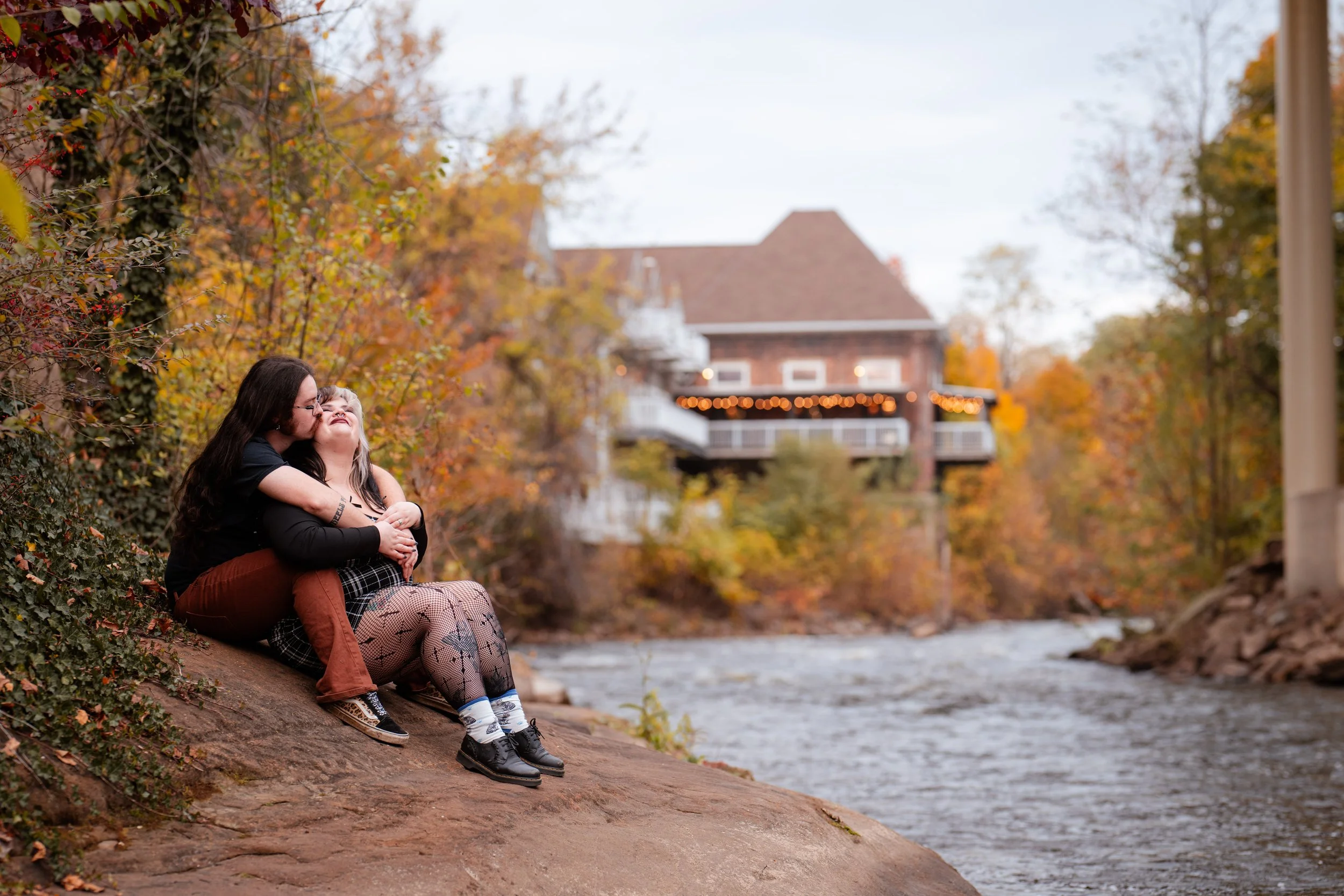 A woman and girl sitting on a rock by a river with autumn trees in the background. The woman is hugging the girl and giving her a kiss on the cheek, with a house decorated with string lights in the background.