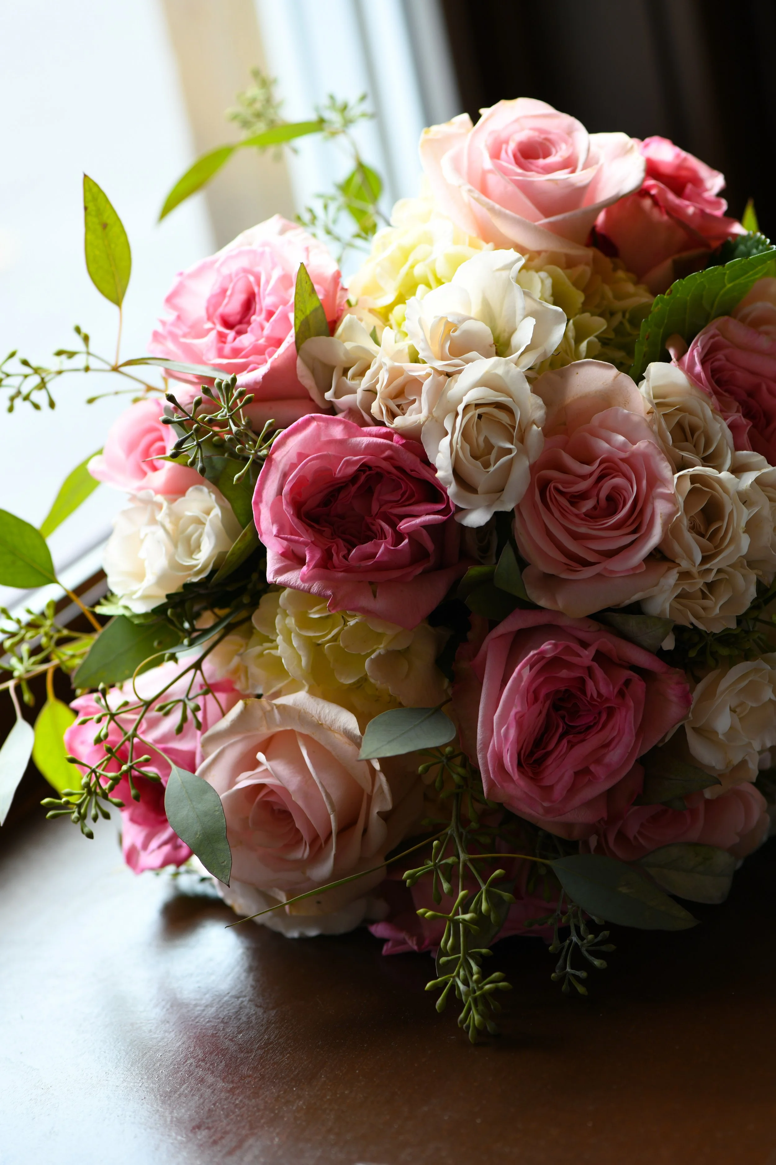 A bouquet of pink and white roses with green leaves on a dark wooden surface, near a window.